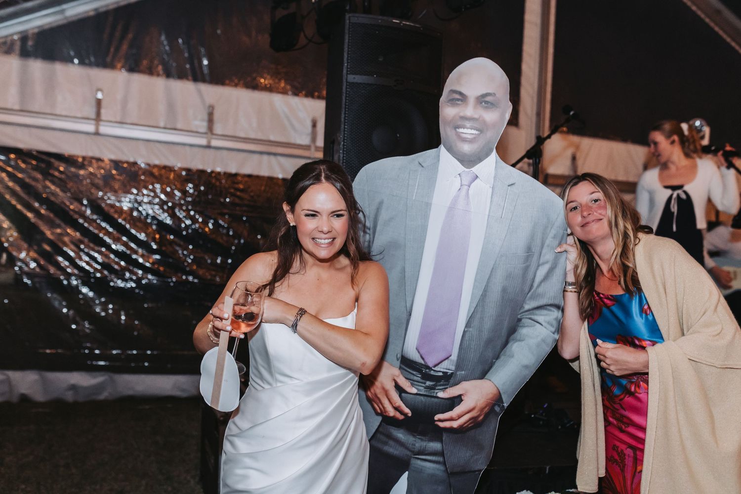 Smiling bride poses with cardboard cutout of celebrity Charles Barkley at outdoor wedding reception.
