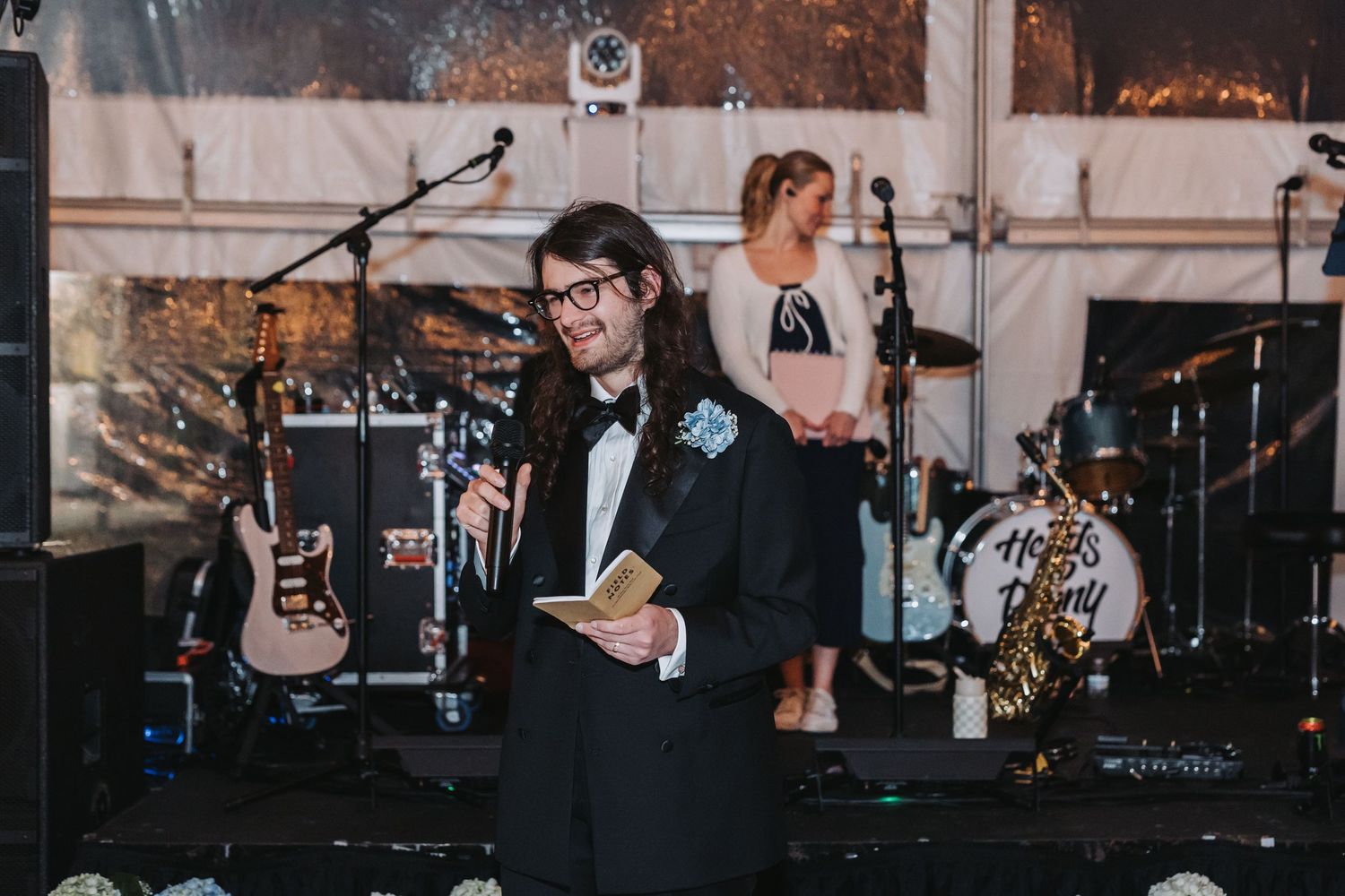 Man in tuxedo with boutonniere speaking at wedding reception podium with live band in background.