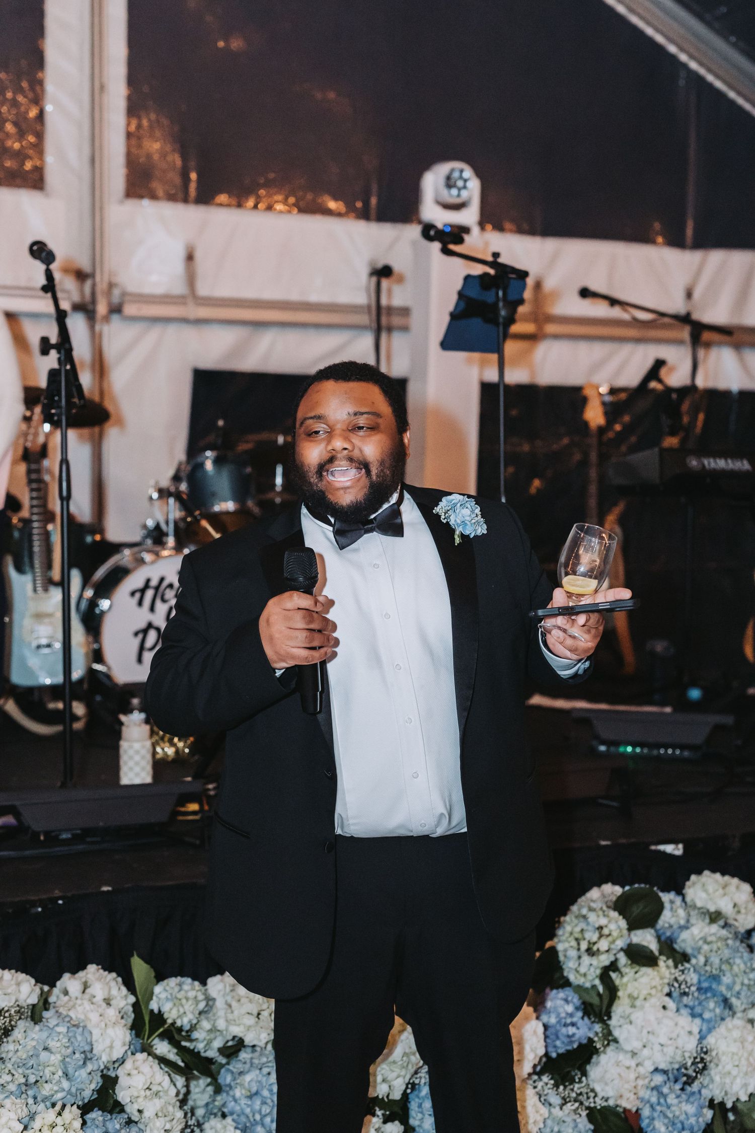 Smiling man in tuxedo with bow tie giving a wedding toast speech while holding a microphone and glass.