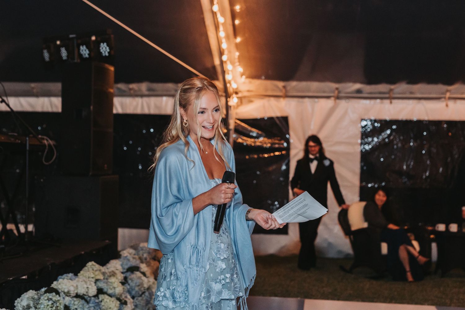 Woman in blue sequin outfit giving a heartfelt speech at a wedding reception inside a tented venue.