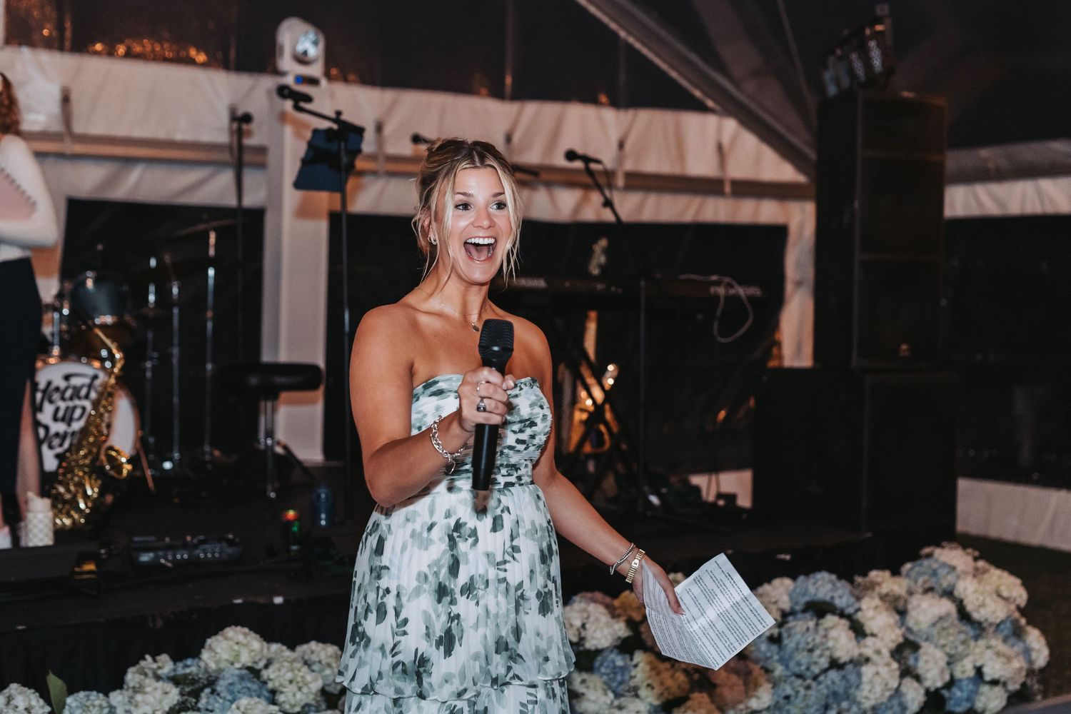 Bridesmaid in floral dress giving a joyful speech with microphone at wedding reception with band.
