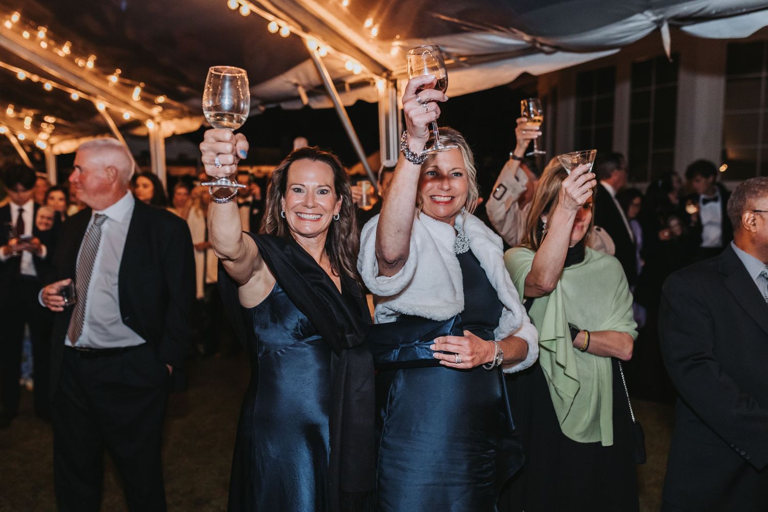 Two women raise glasses in a toast at a festive wedding reception under string lights.