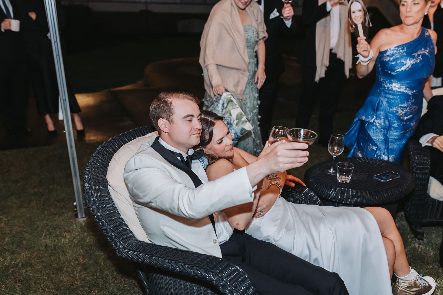 Bride and groom toasting drinks while seated in wicker chairs at outdoor wedding reception at night.