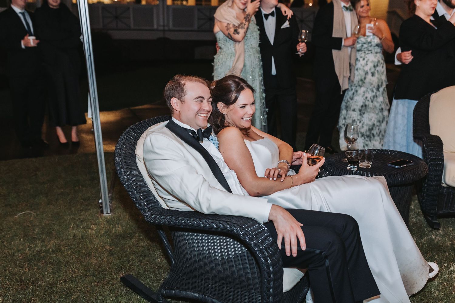 Bride and groom relaxing together in wicker chairs outdoors during evening wedding reception celebration.