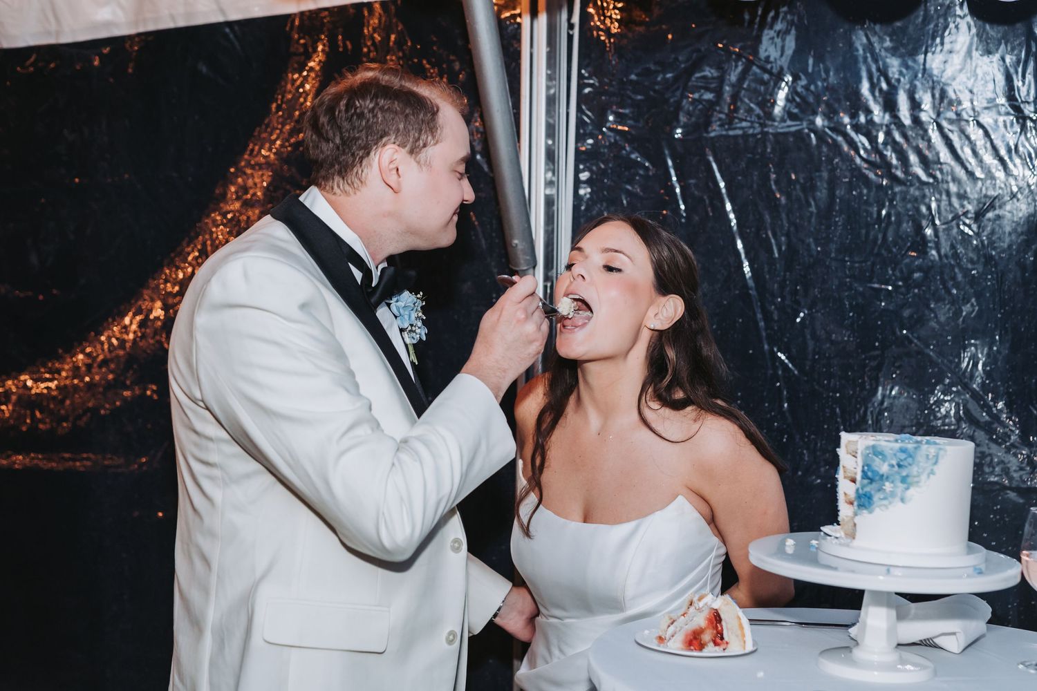 Groom feeds bride a slice of wedding cake at elegant white-tented reception.