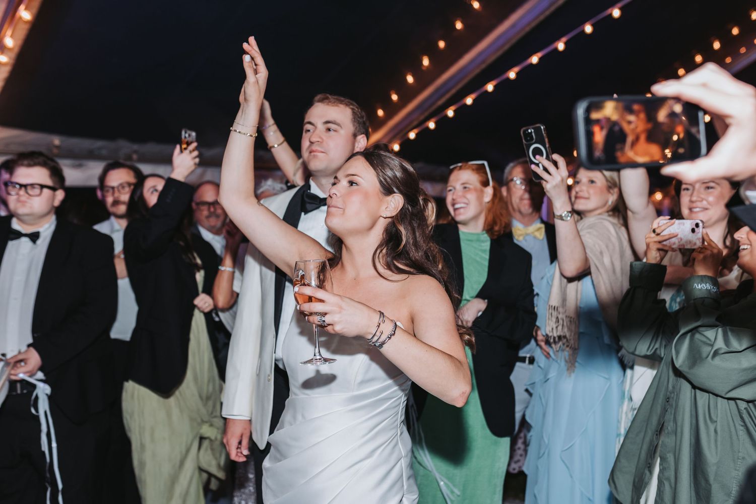 Bride in white gown celebrating with groom and guests raising glasses at lively wedding reception.