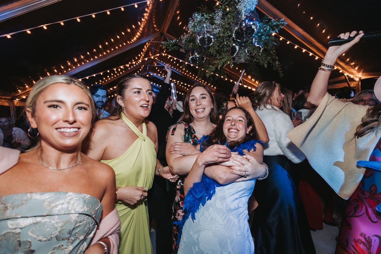Excited wedding guests reach for bouquet toss under string lights at lively outdoor reception celebration.