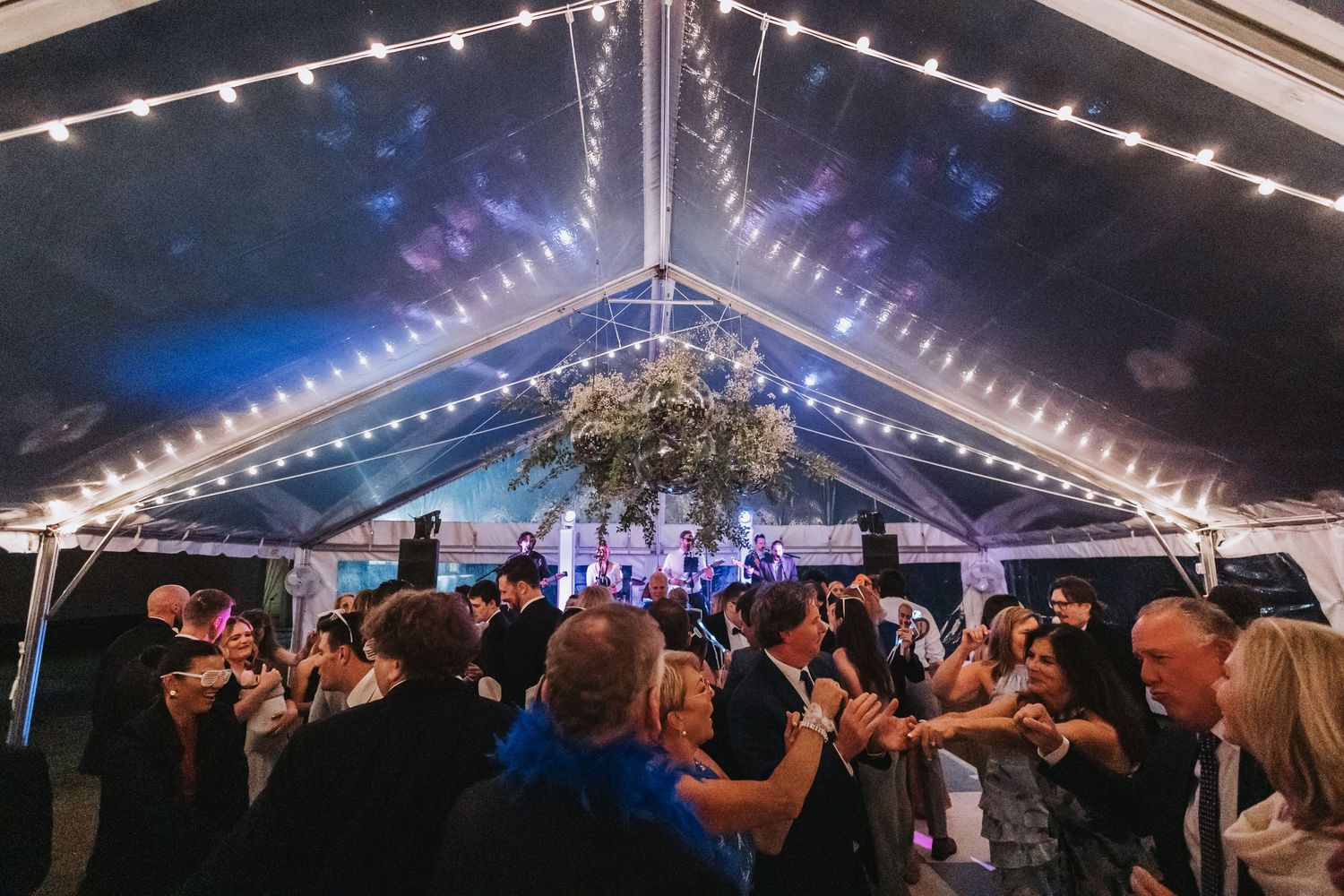 Guests dancing under string lights inside a decorated clear wedding reception tent.