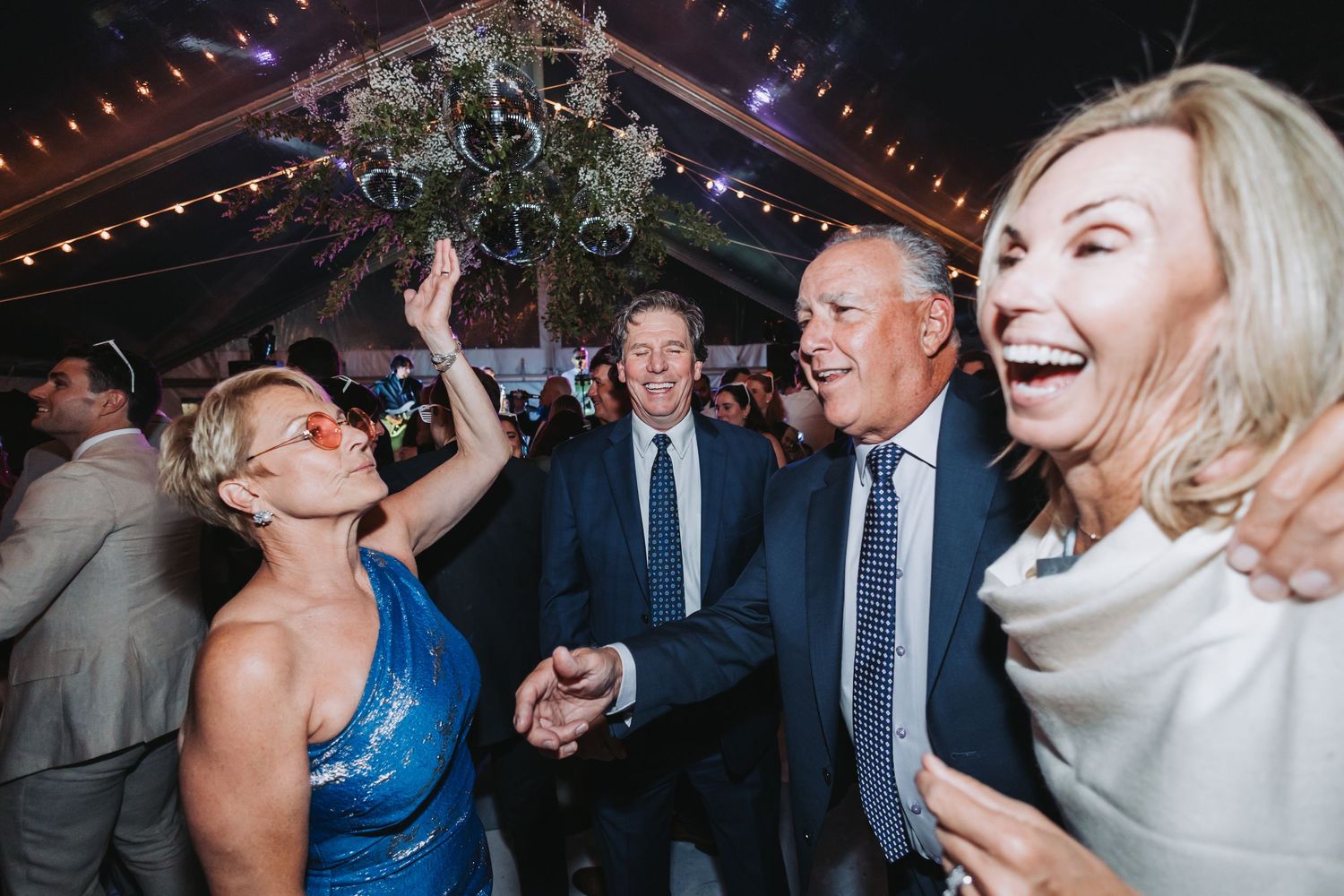 Joyful wedding guests dancing and laughing together under a decorated tent at evening reception.