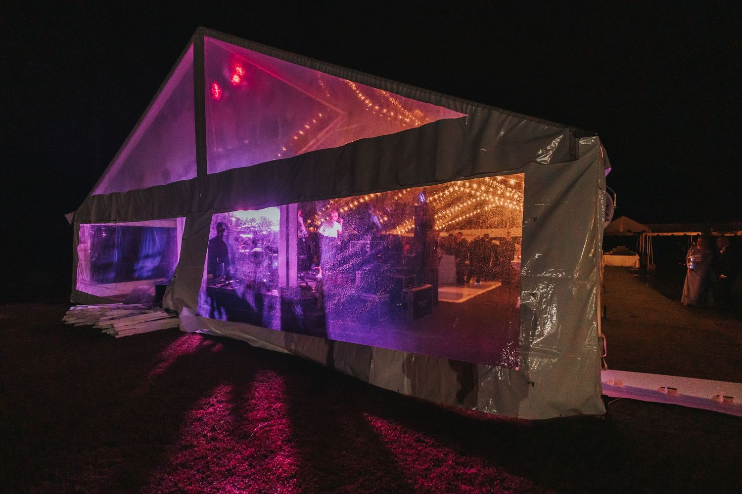 Clear wedding tent glows with purple and pink lights at night during outdoor reception celebration.