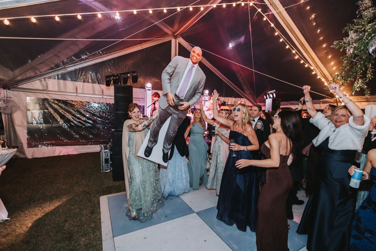 Groomsman lifted by wedding guests on checkered dance floor during lively outdoor reception celebration at night.