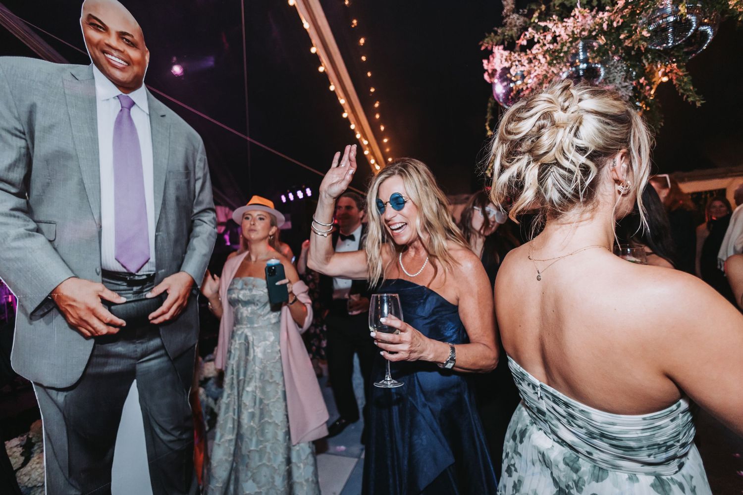 Wedding reception guests dancing with wine glasses under string lights in elegant tent venue.