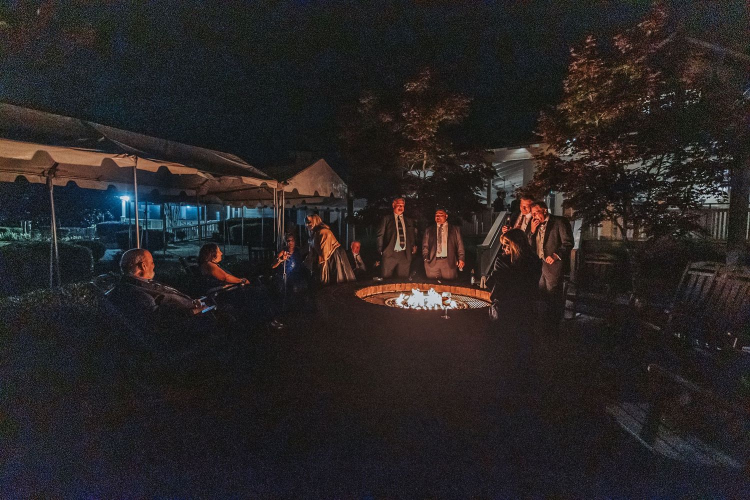 Wedding guests gather around glowing fire pit outdoors at night near white reception tent.