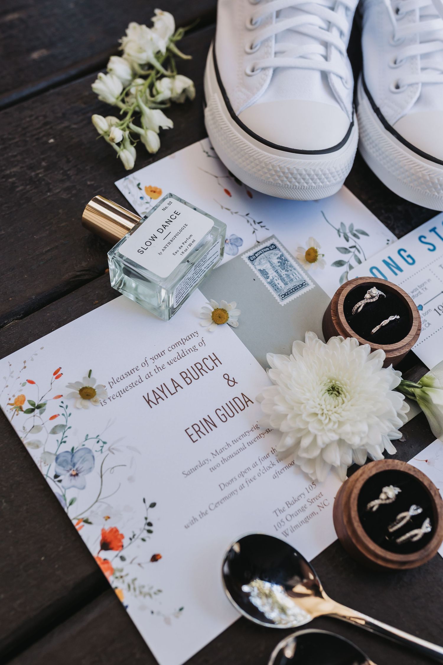Wedding flat lay with floral invitation, white sneakers, perfume, rings, and flowers on dark wood.