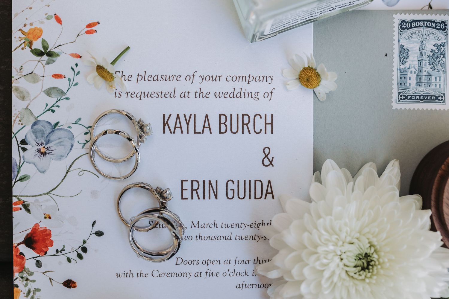 Wedding invitation for Kayla Burch and Erin Guida with floral design, gold rings, and white daisy on elegant stationery.