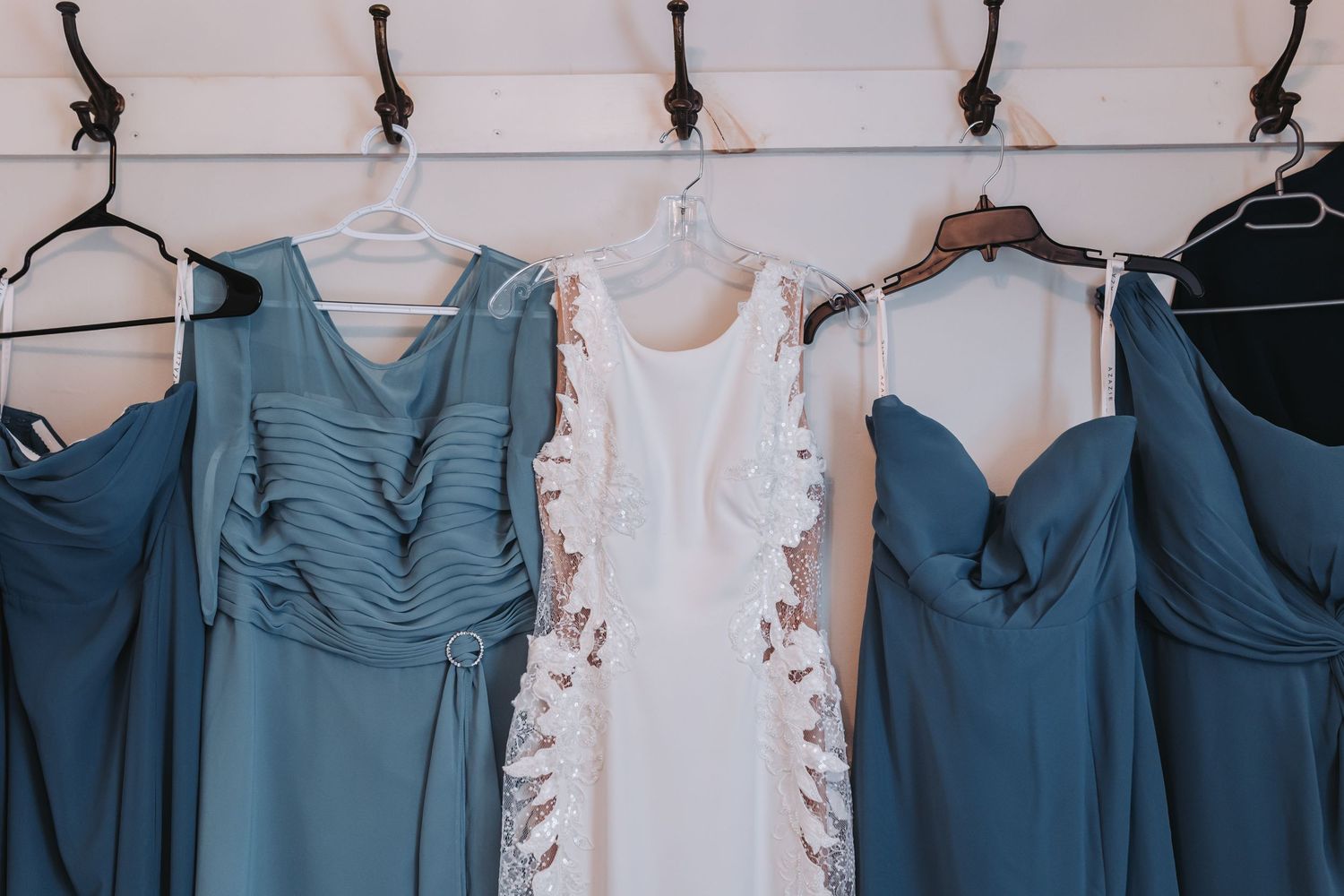 White wedding dress flanked by dusty blue bridesmaid gowns hanging on hooks against a white wall.