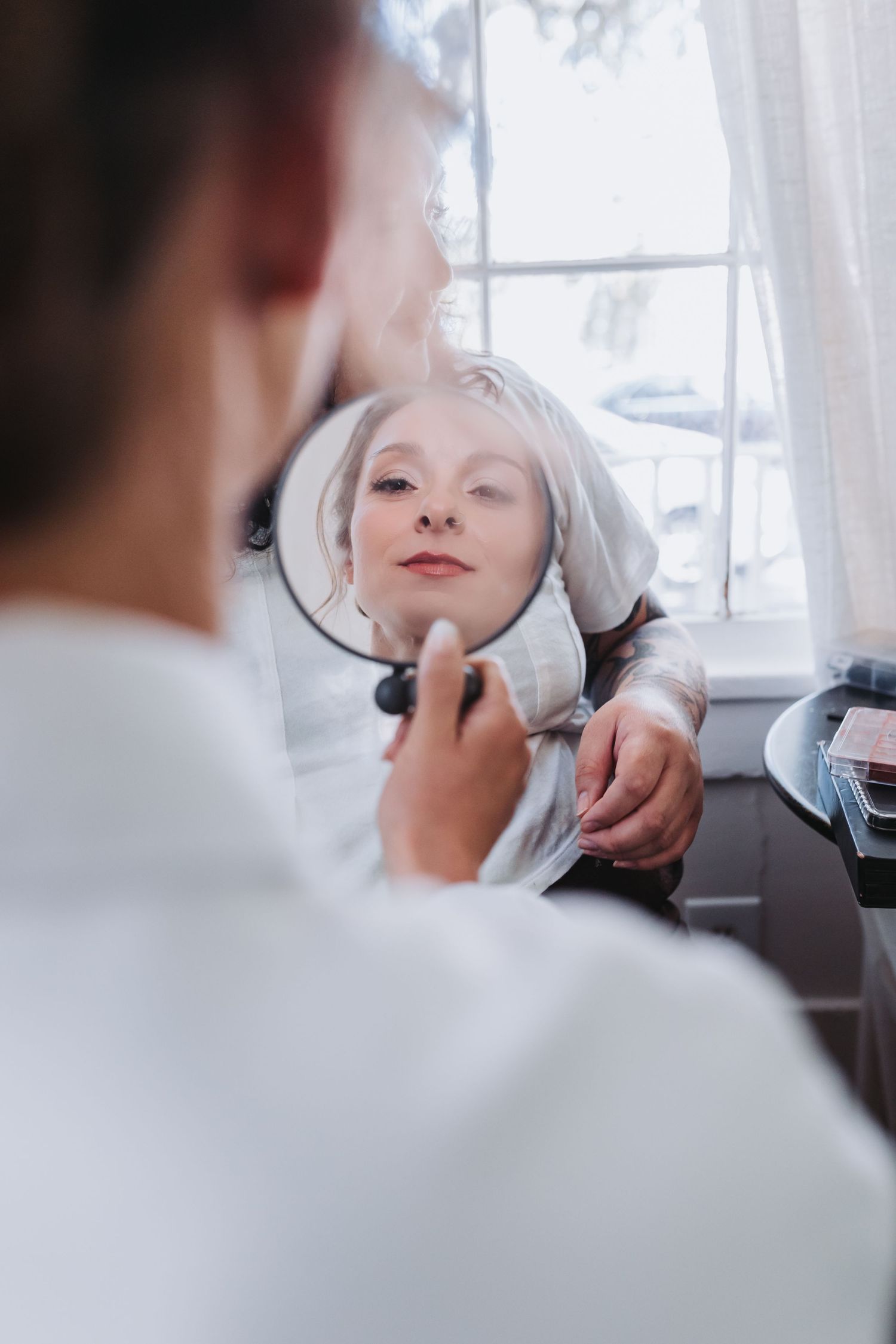 Bride applying makeup while looking into a round mirror during wedding day preparations.