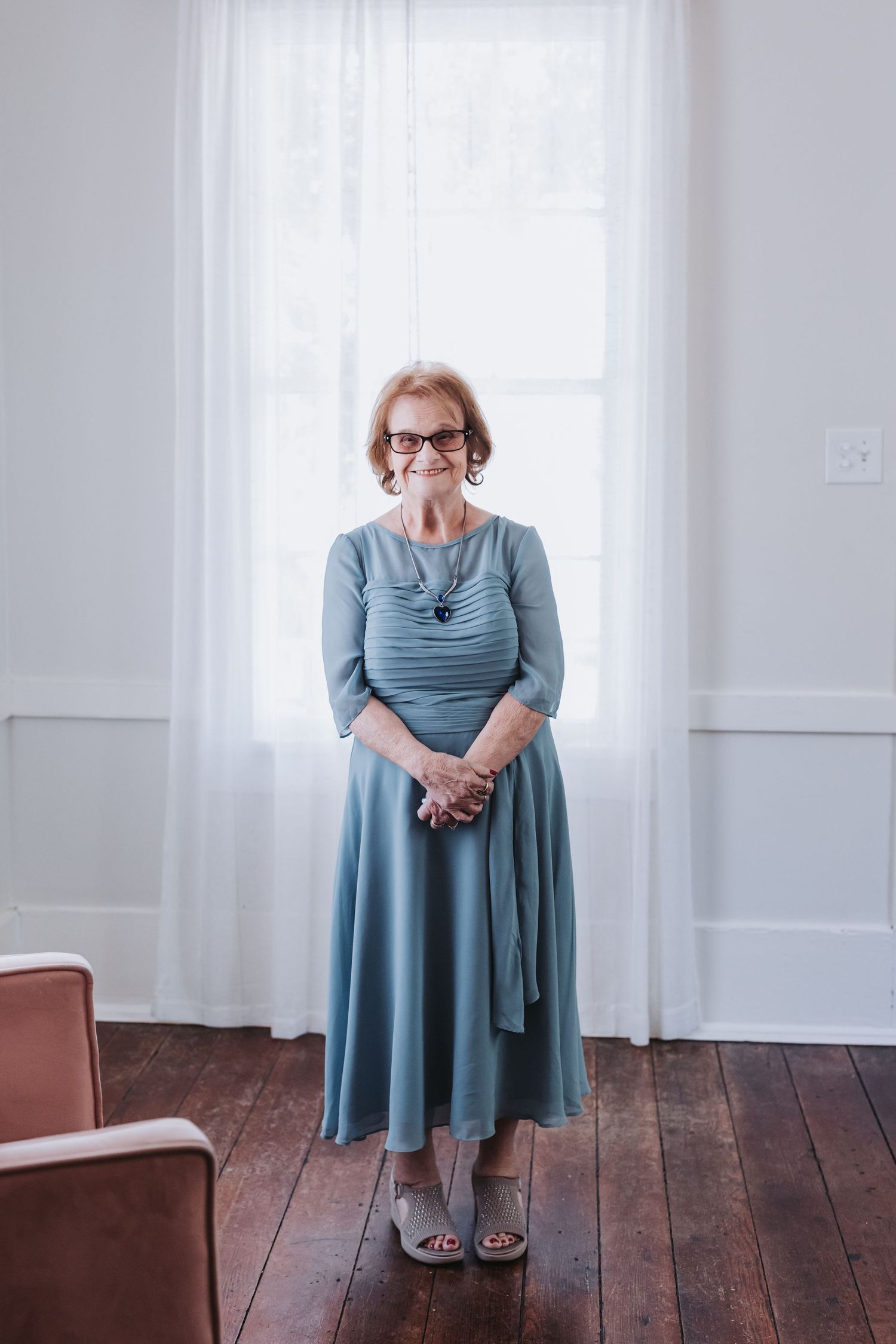 Elegant senior woman in dusty blue midi dress with necklace and glasses standing by bright window on hardwood floor.