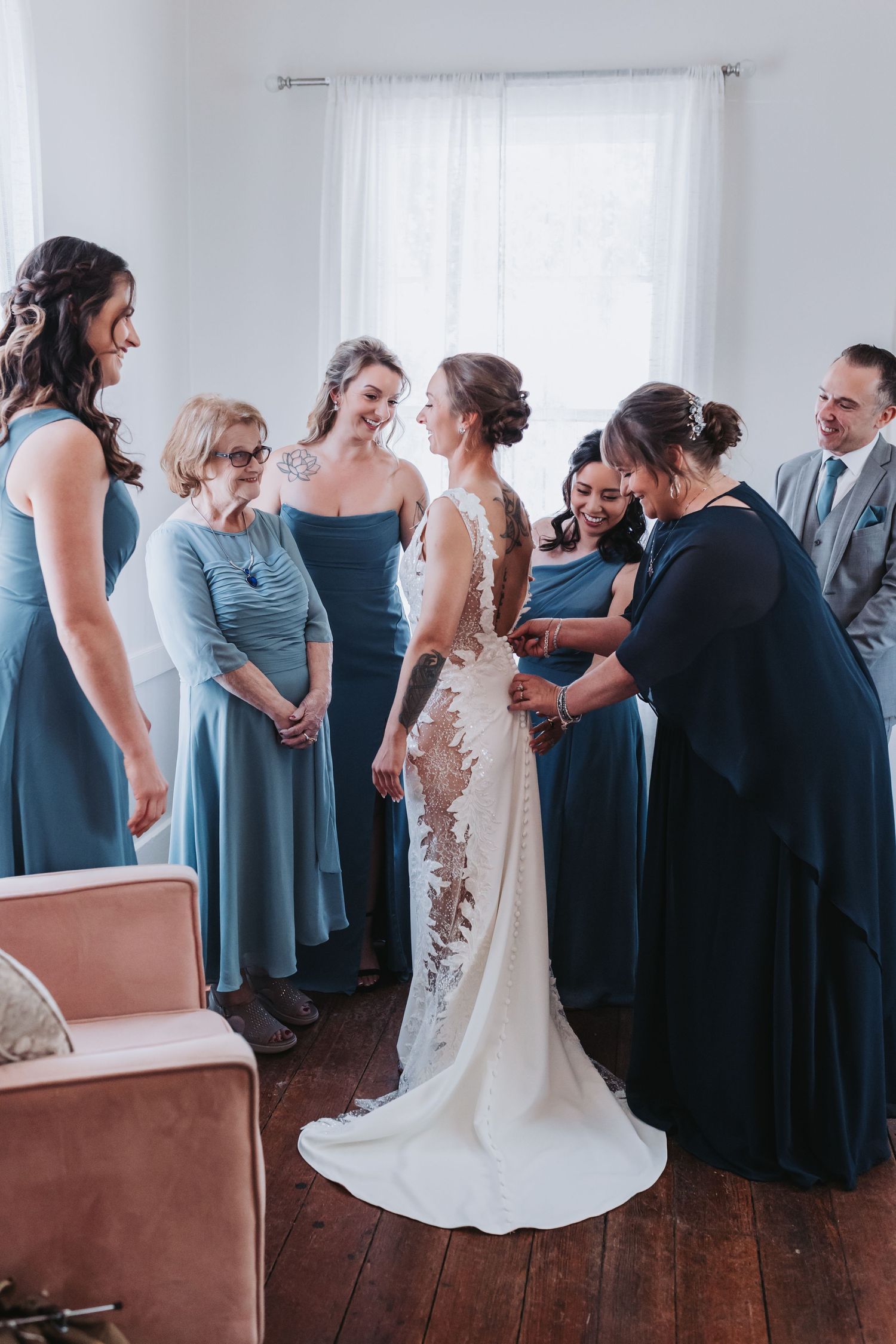 Bridesmaids in dusty blue dresses helping bride into her lace wedding gown in a bright room.