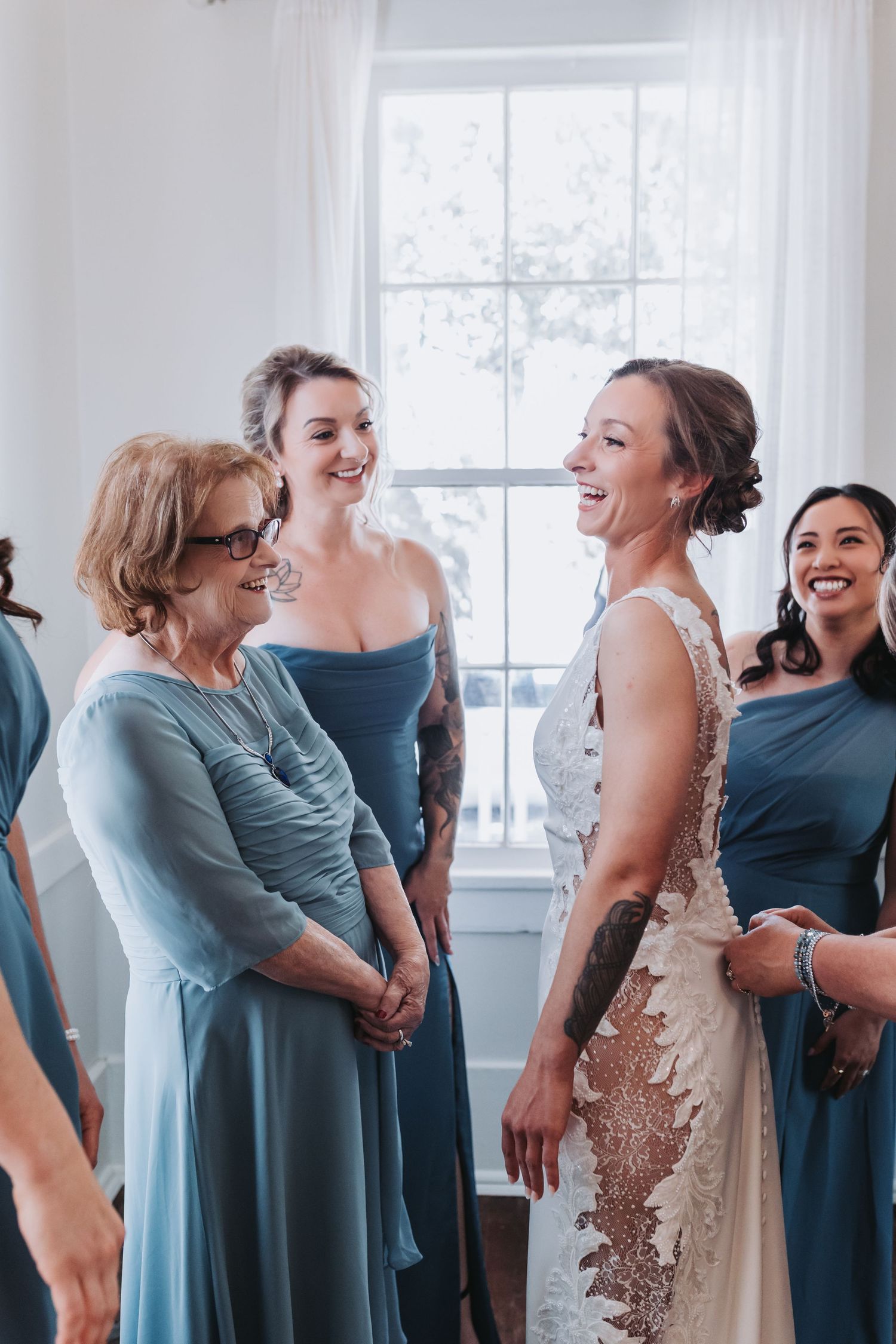 Bride laughing with bridesmaids and mother in blue dresses during wedding preparations.
