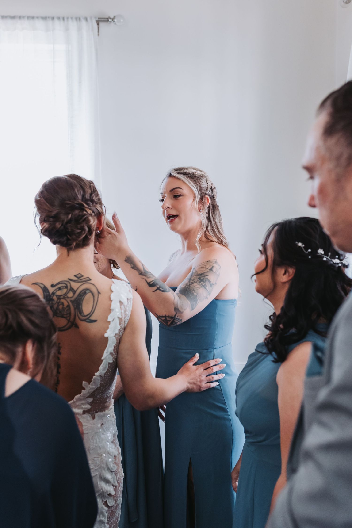 Bridesmaids in blue dresses help a tattooed bride with her earring during wedding preparations indoors.