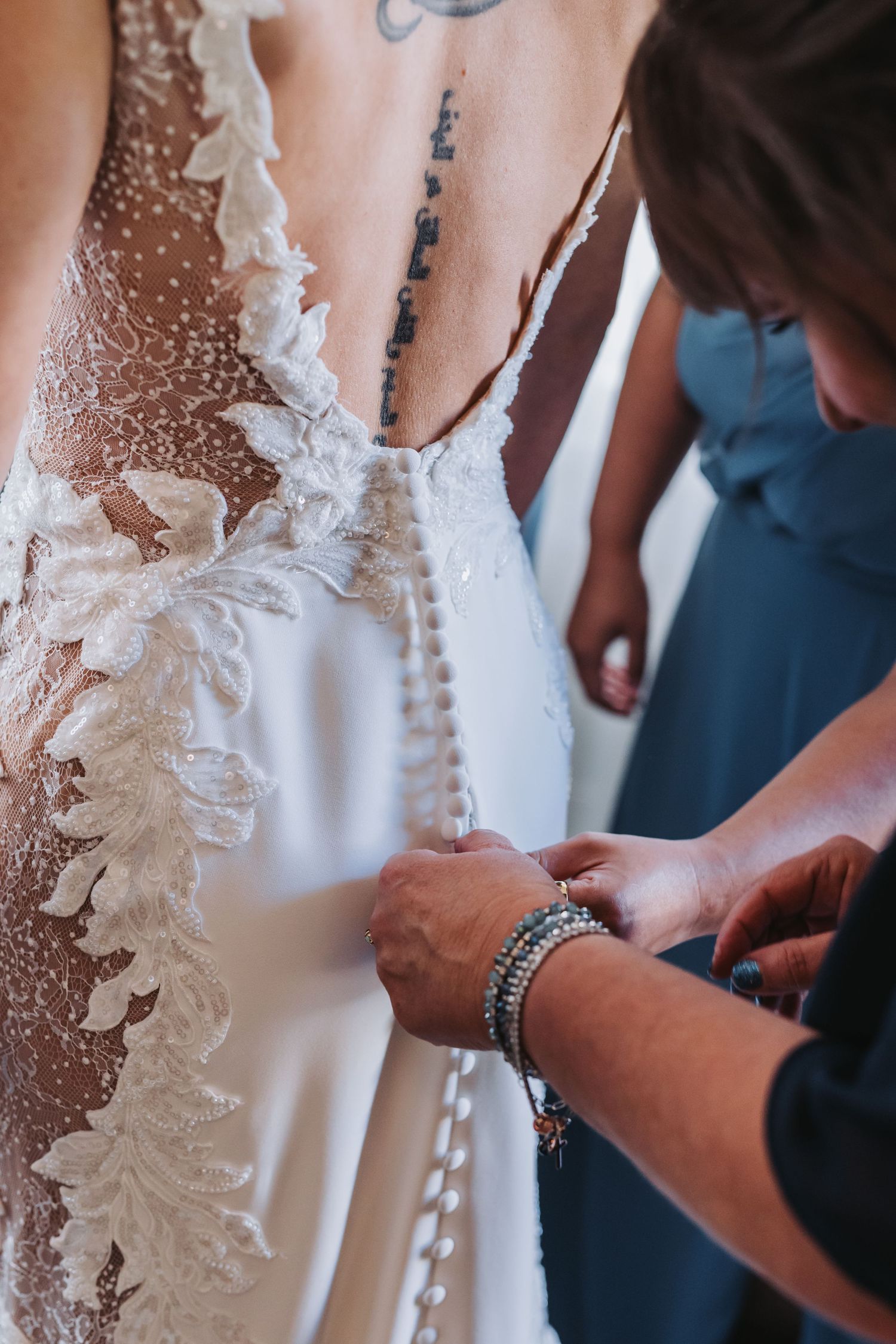 Close-up of bridesmaids fastening pearl buttons on the back of a lace wedding gown.