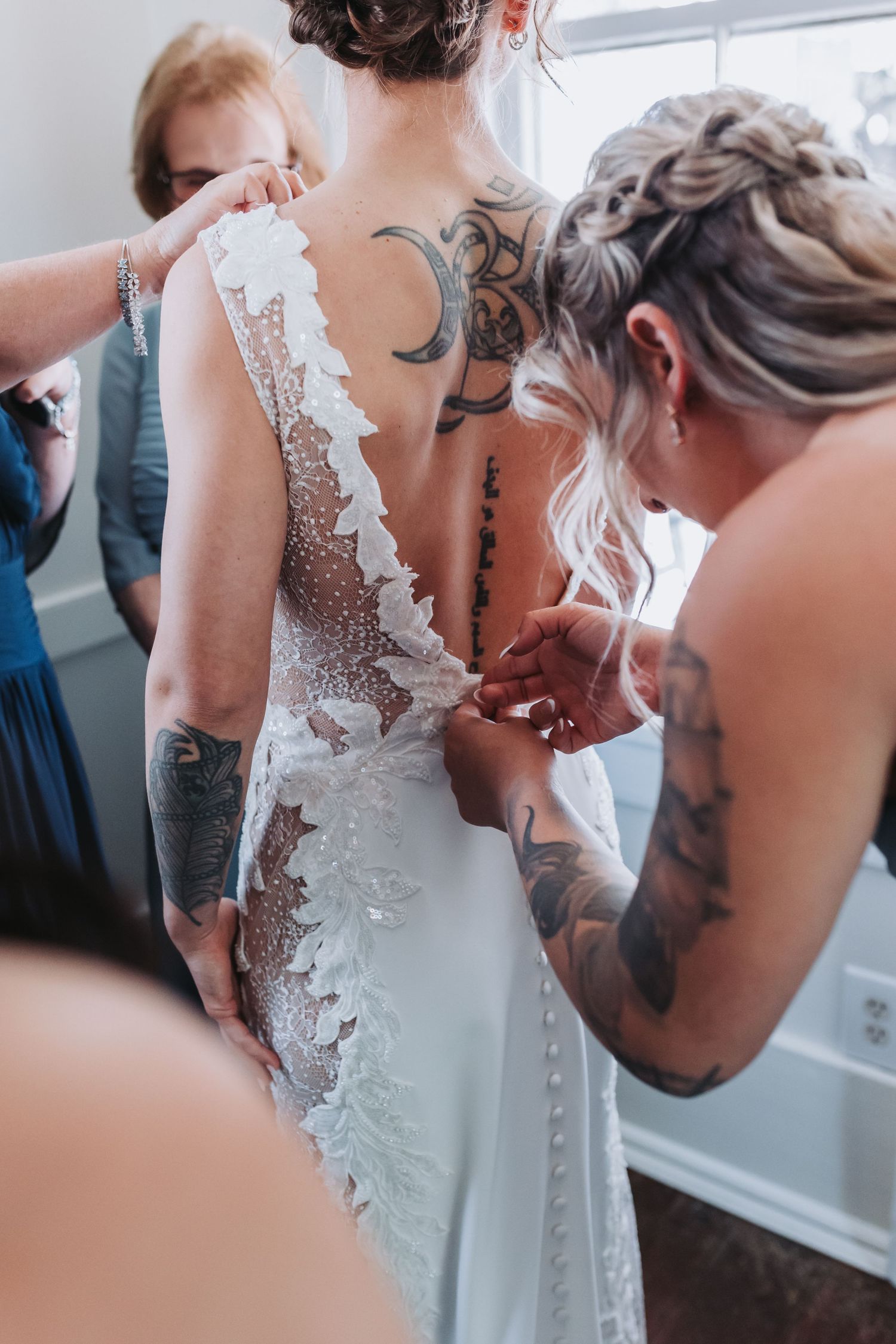 Bridesmaids helping bride button up lace wedding dress showing back tattoos.