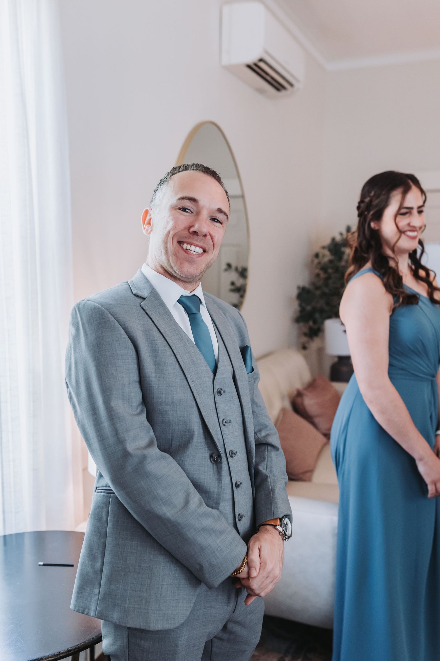 Smiling groom in a grey three-piece suit standing with a bridesmaid in a blue dress.