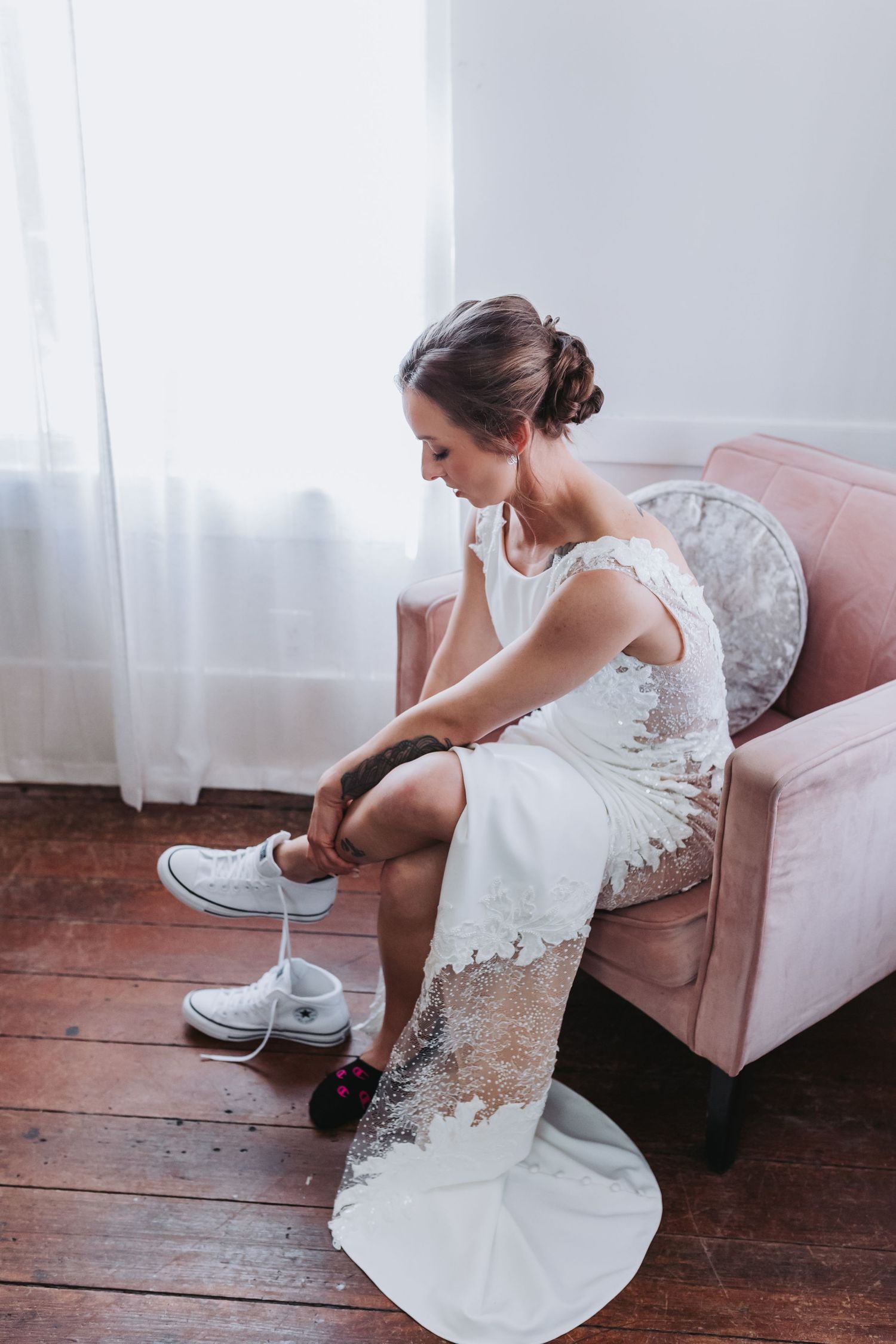 Bride in a lace wedding dress sitting on a pink chair putting on white Converse sneakers.