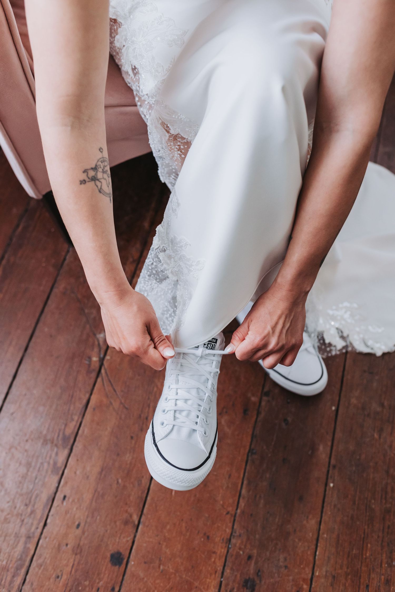 Bride in white wedding dress tying white Converse sneakers on rustic hardwood floor.