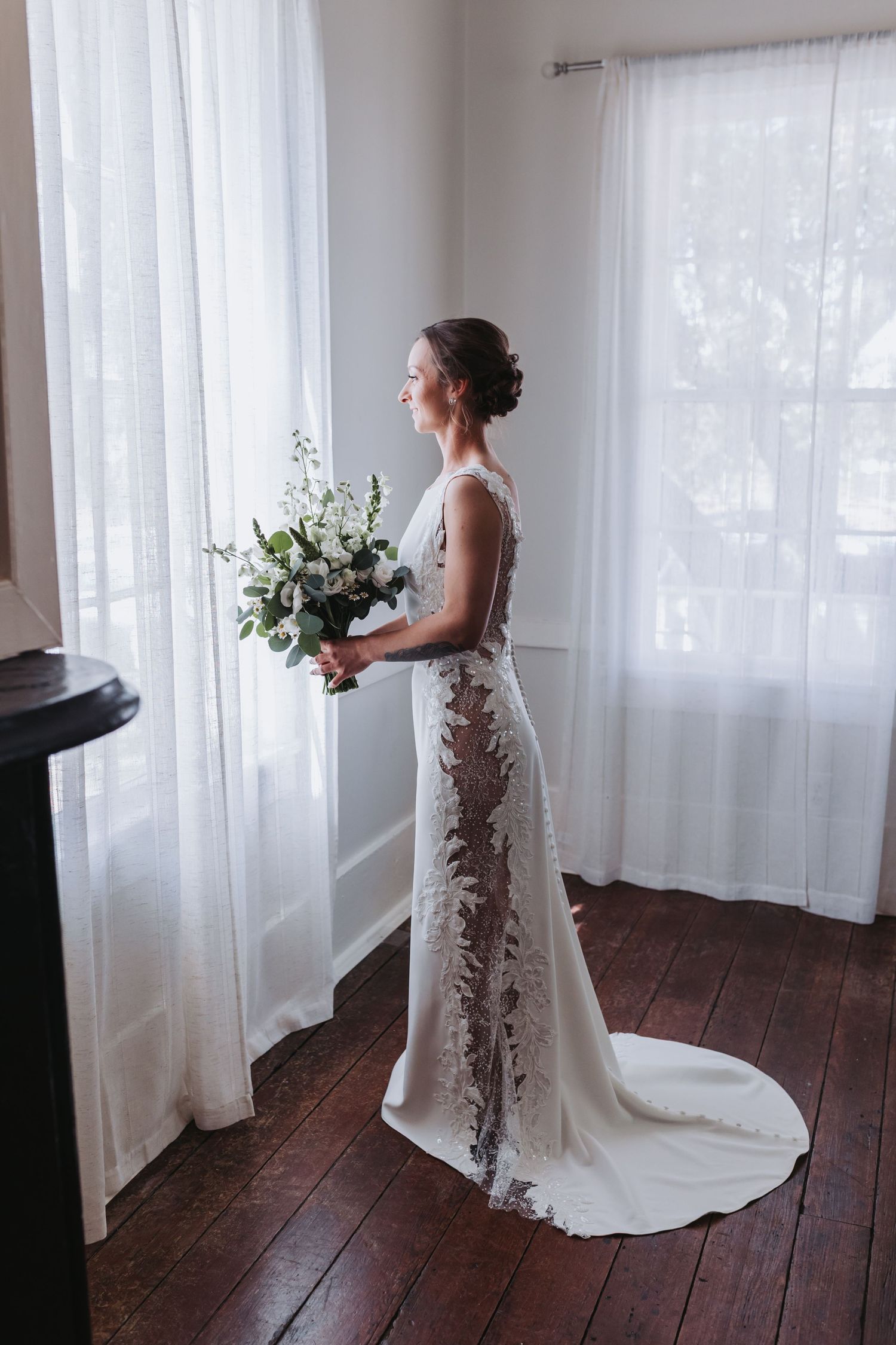Bride in lace wedding gown with train holds white floral bouquet while gazing out sunlit window.
