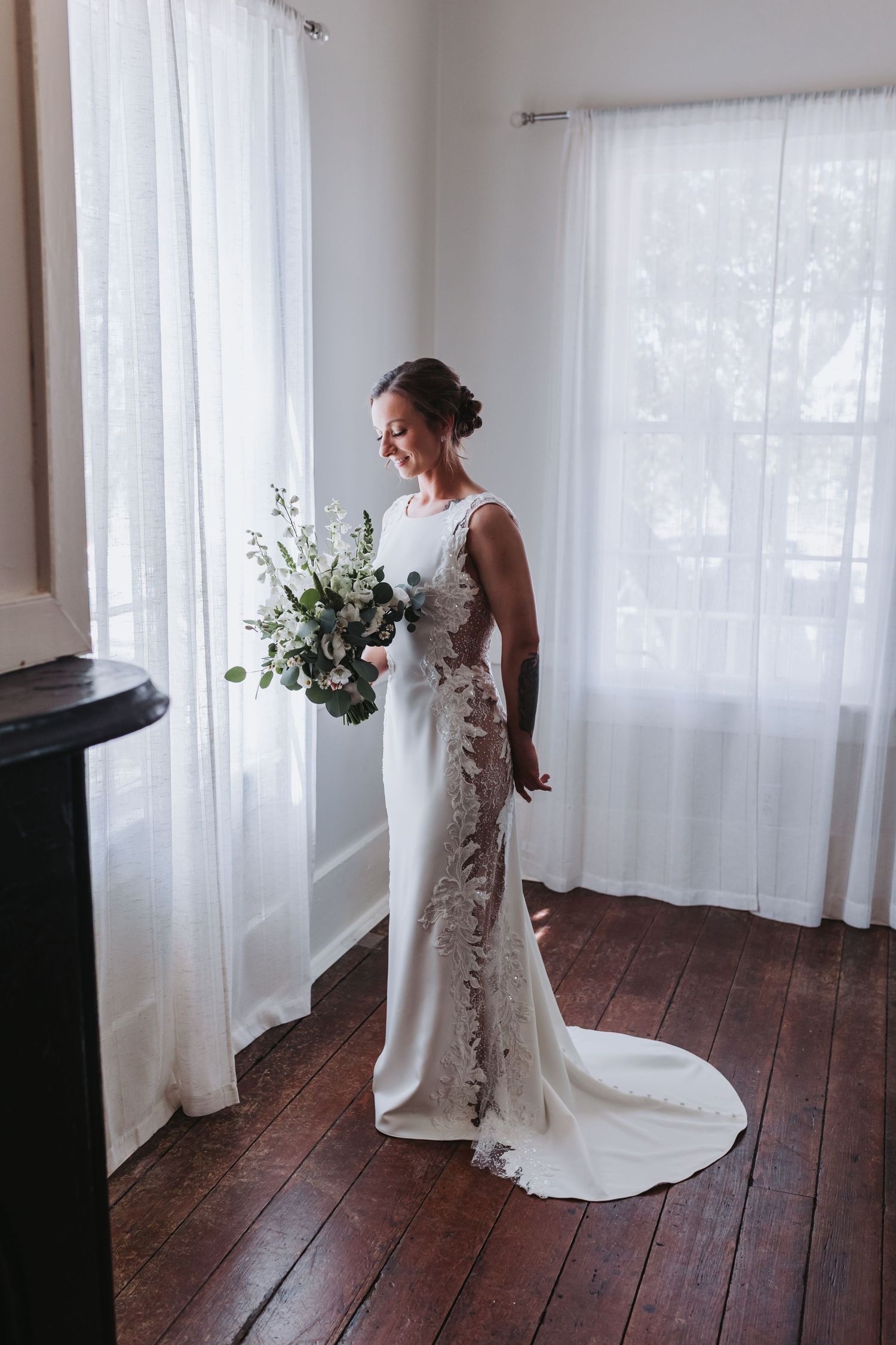 Bride in a fitted lace wedding gown holding a white floral bouquet by a sunlit window.