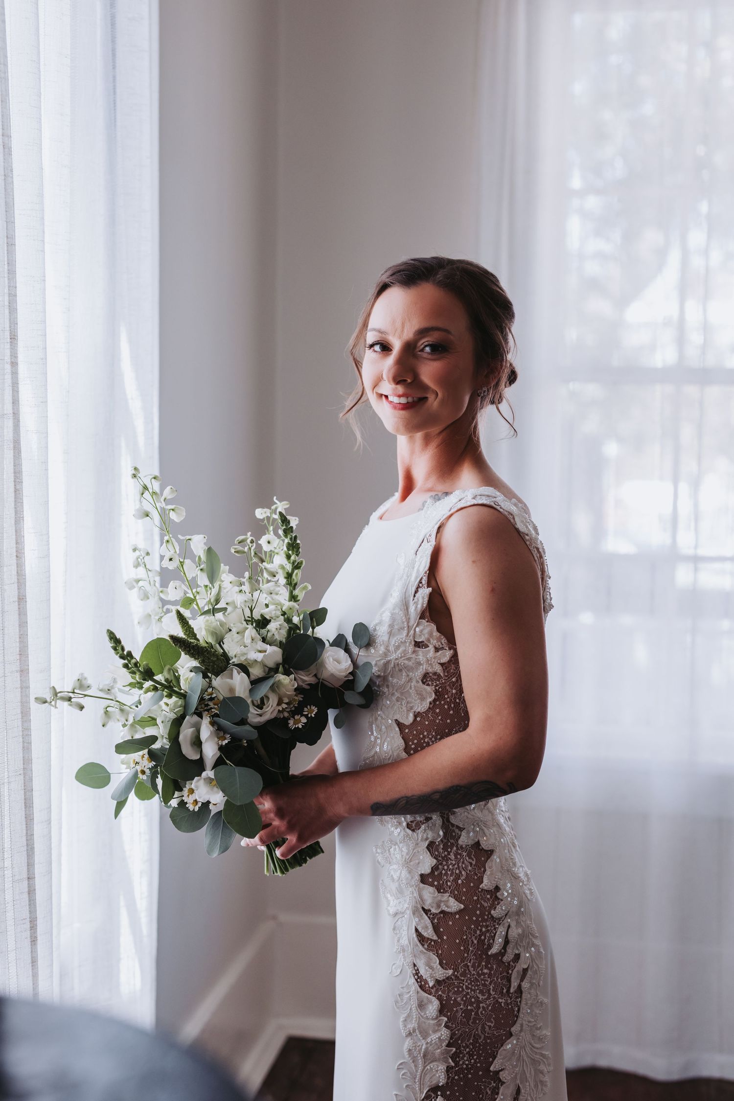 Smiling bride in lace wedding dress holds white and green bouquet while standing near bright window.