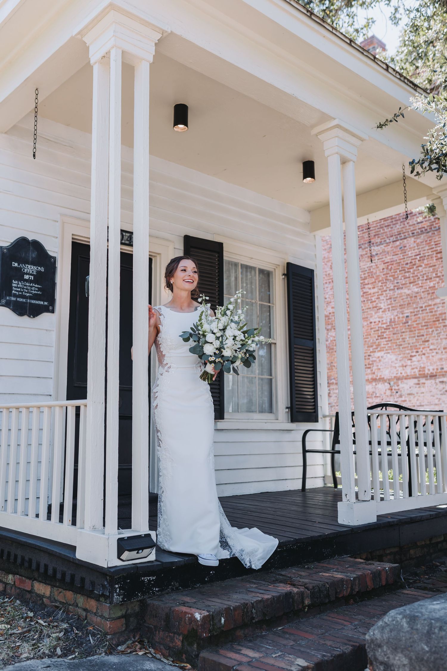 Smiling bride in white wedding dress holding floral bouquet on porch of white historic building.
