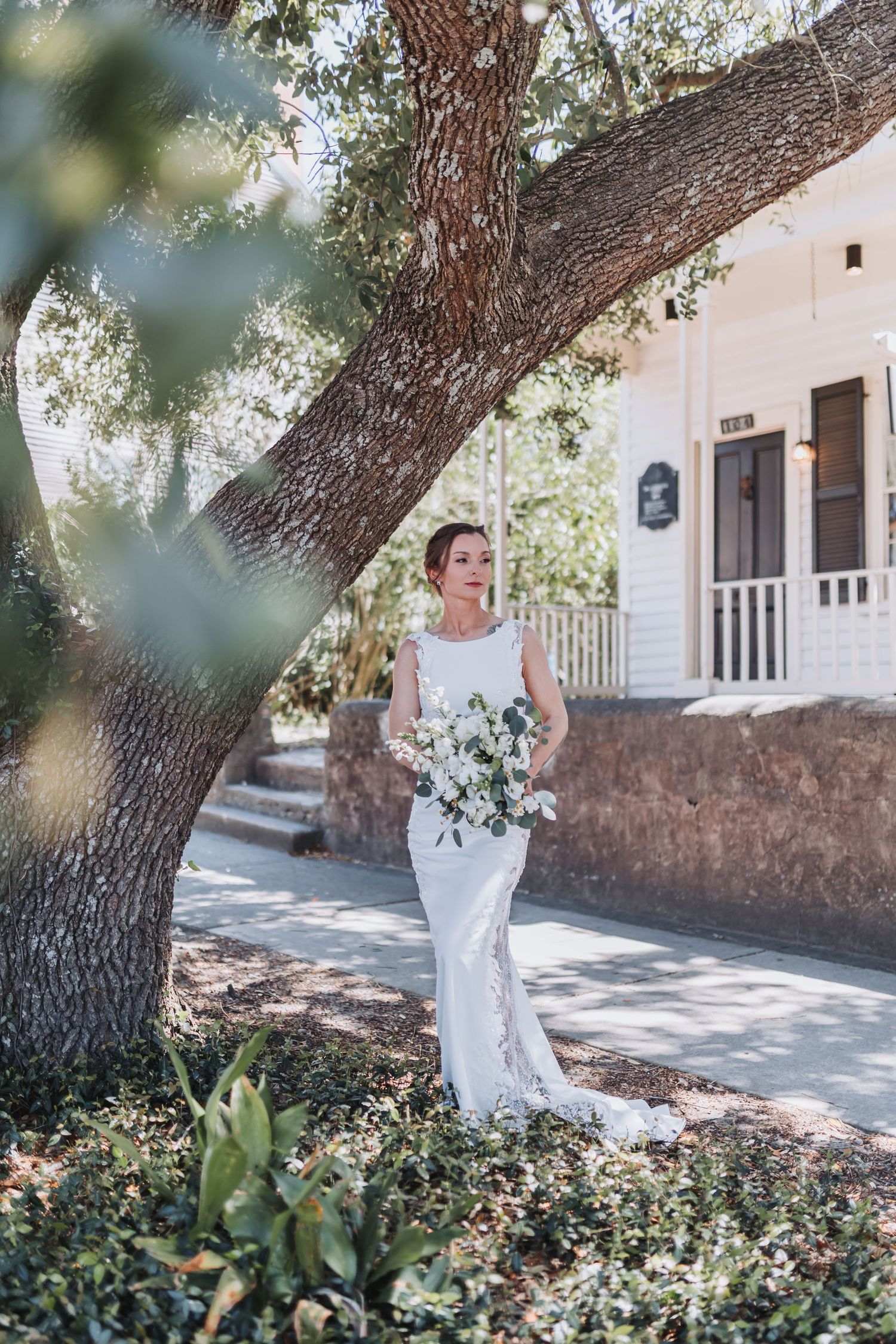 Bride in fitted lace wedding gown with bouquet stands beneath large oak tree outside white venue.