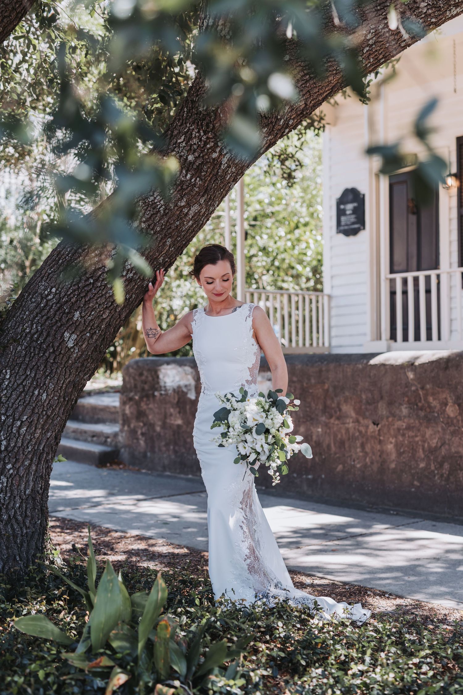 Elegant bride in white gown holds bouquet of white flowers beside a large oak tree outside a historic venue.