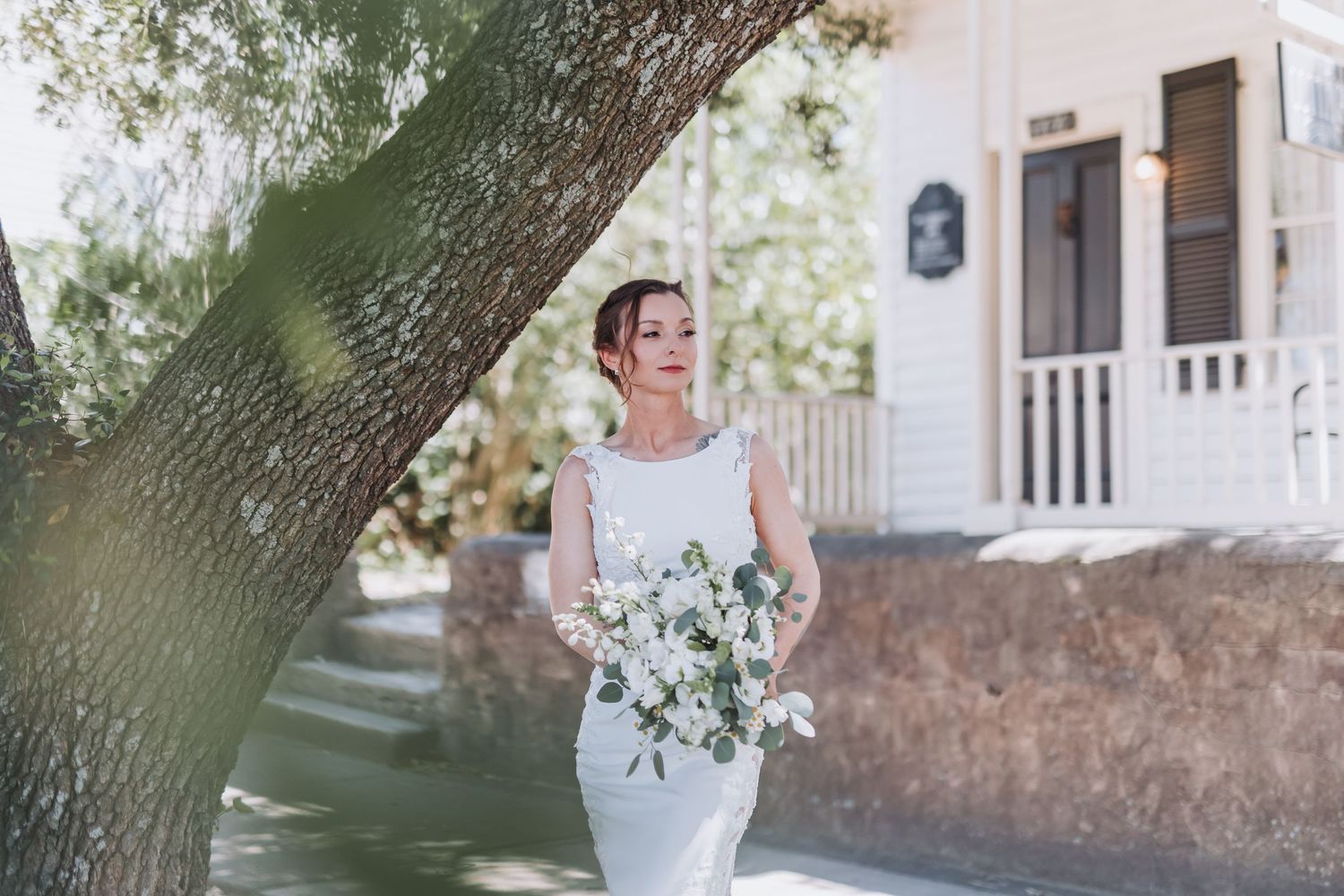 Bride in fitted white gown holding bouquet standing near large tree outside white venue.