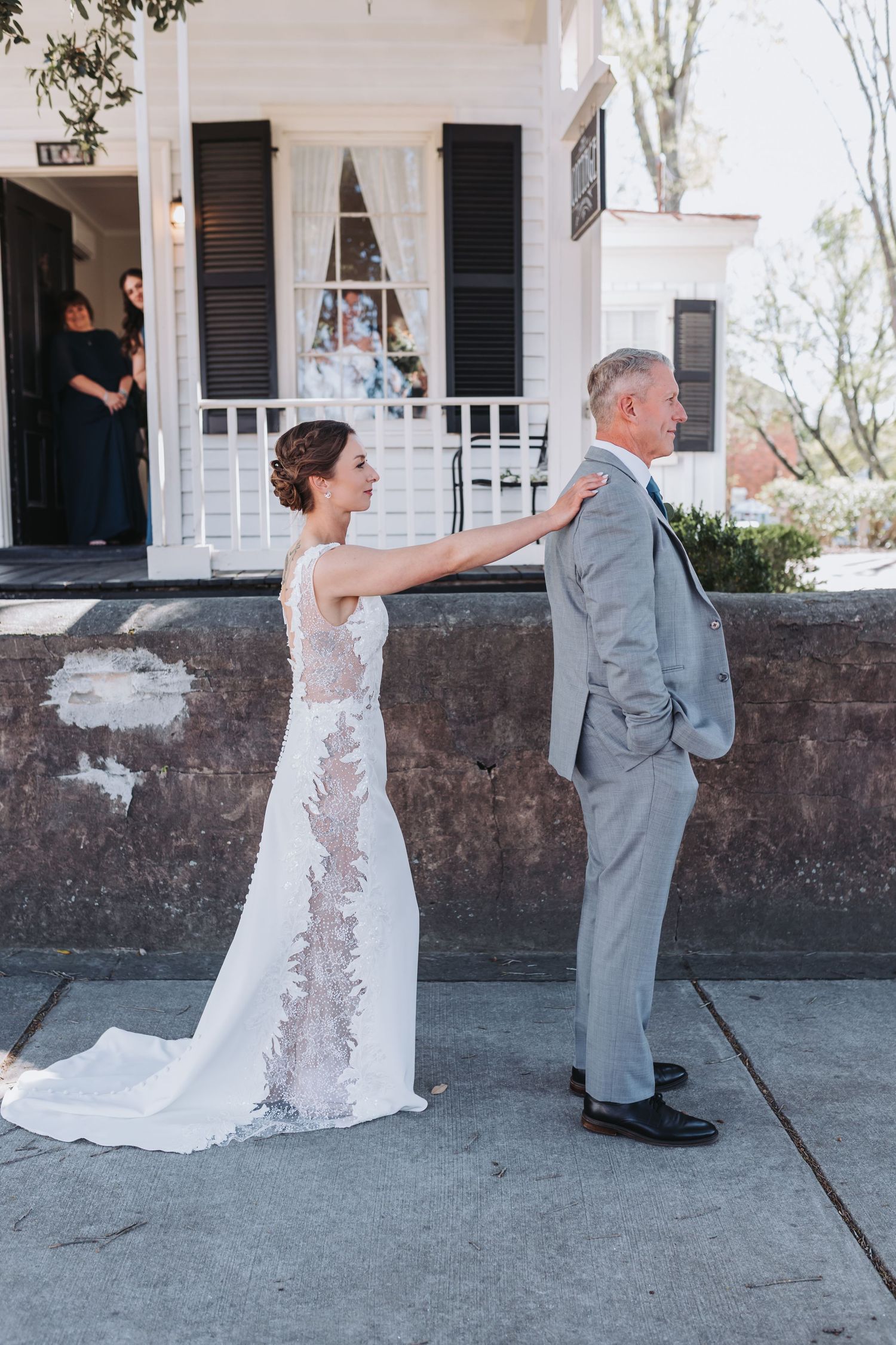 Bride in lace wedding gown touching grooms shoulder during outdoor first look moment.