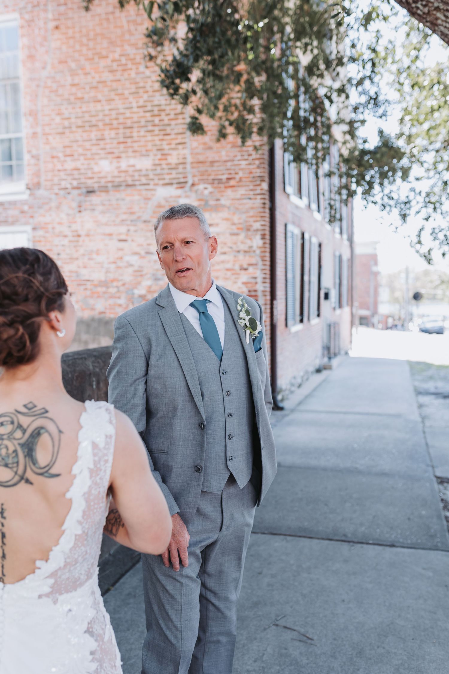 Groom in gray suit with teal tie reacts emotionally during first look with bride in lace wedding dress outdoors.