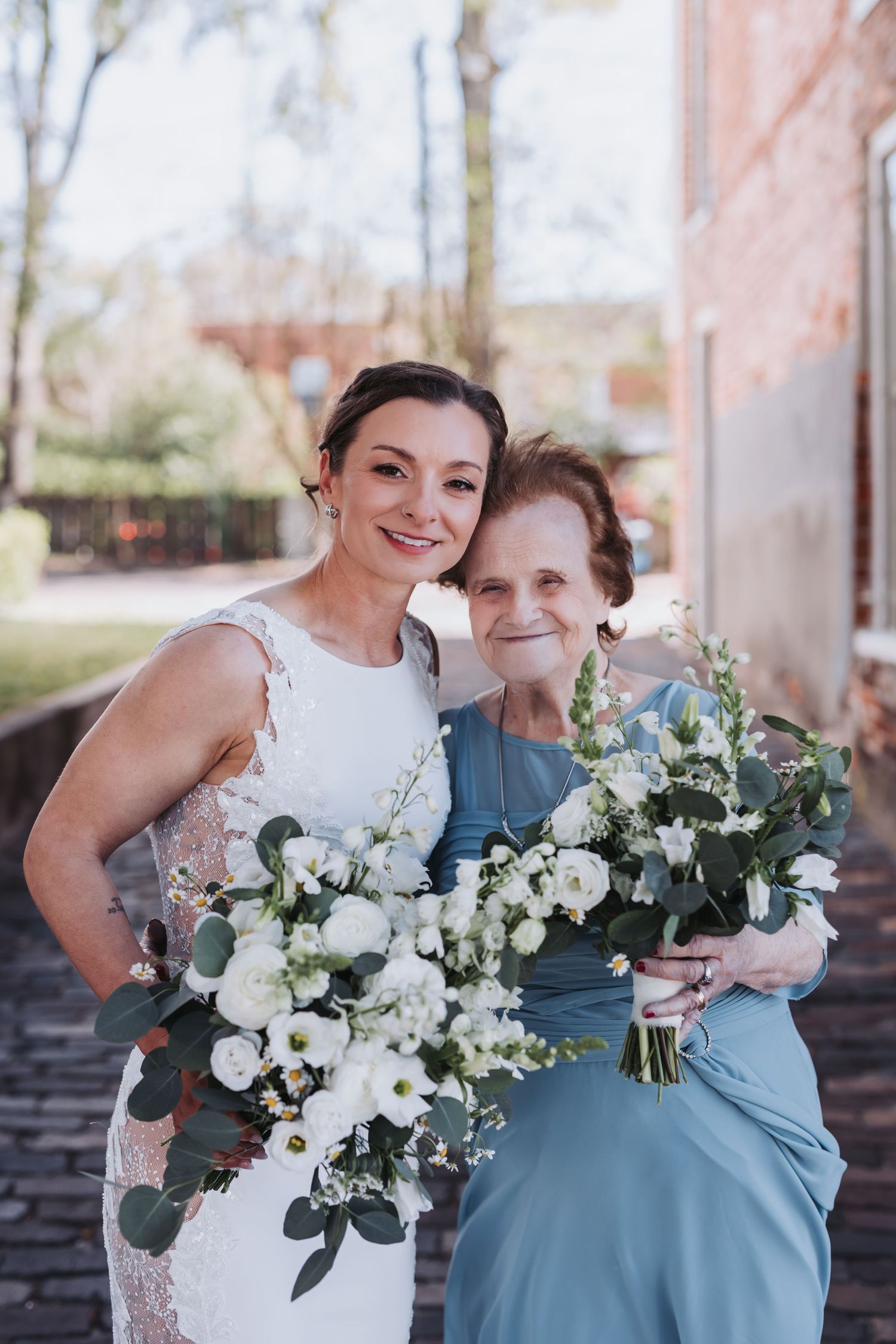 Smiling bride in white lace gown poses with elderly woman in blue dress holding white floral bouquets outdoors.