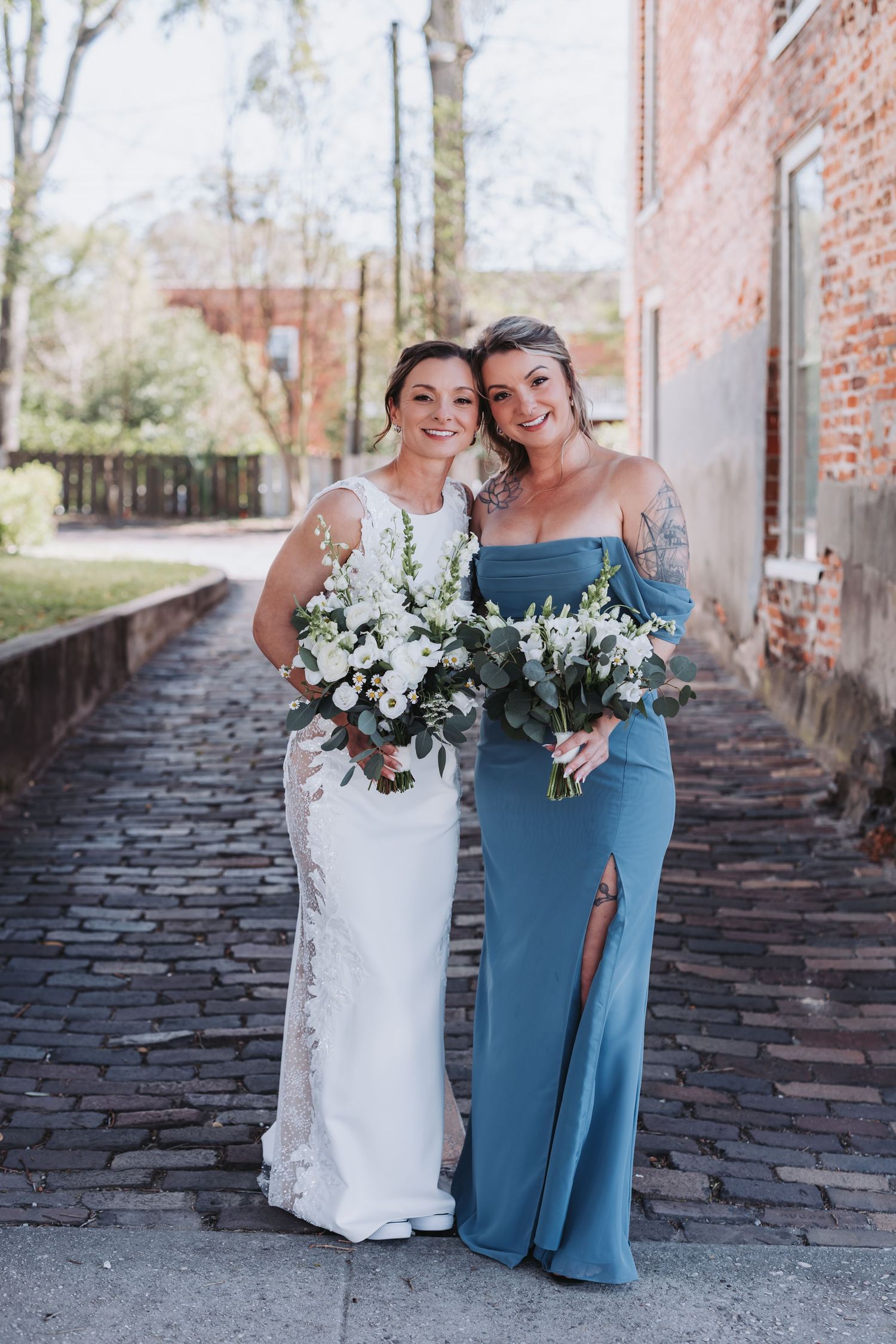 Bride in white lace gown and bridesmaid in blue dress holding white bouquets on cobblestone path.