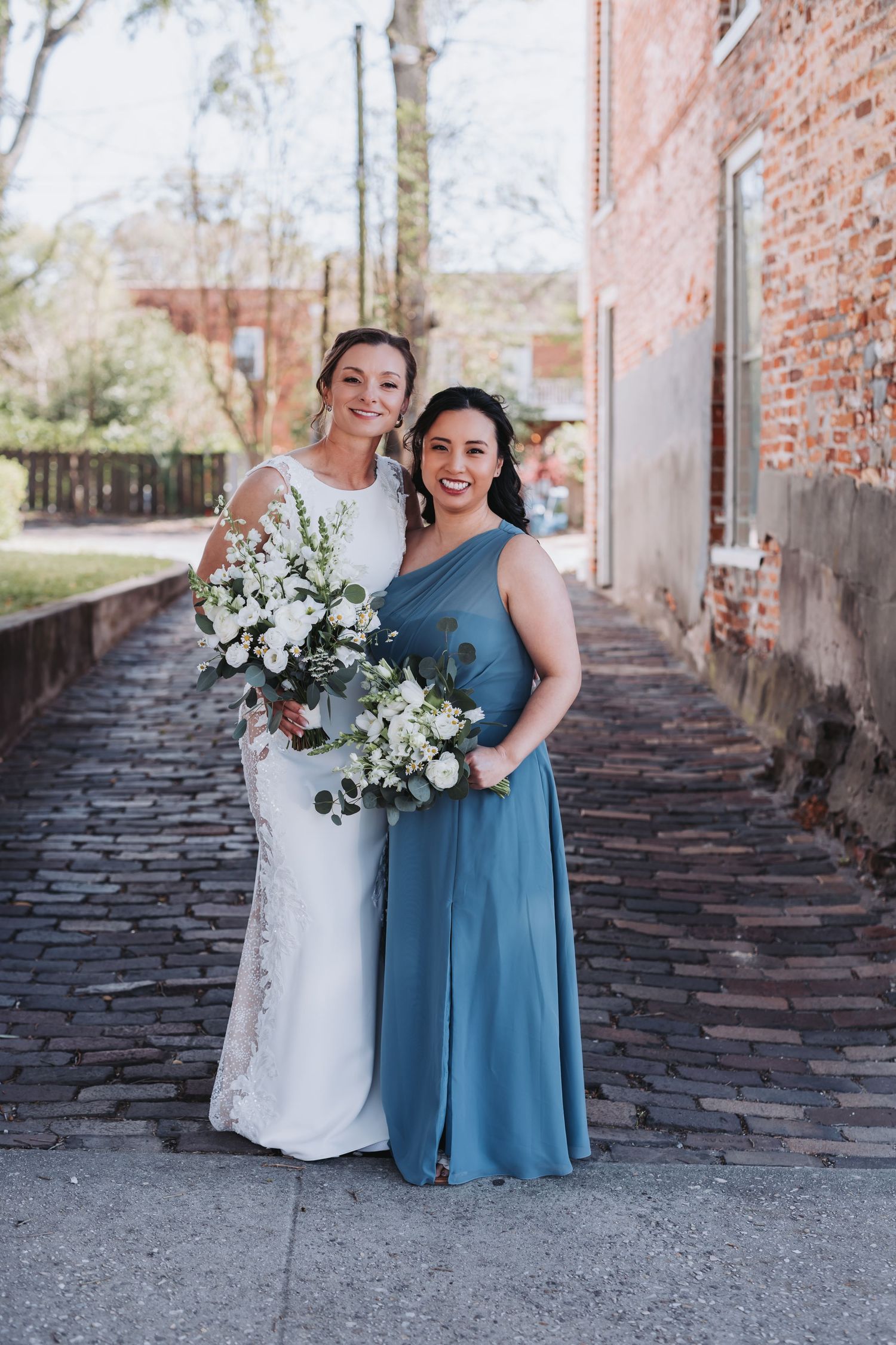 Bride and bridesmaid in blue dress posing with white bouquets on cobblestone path.