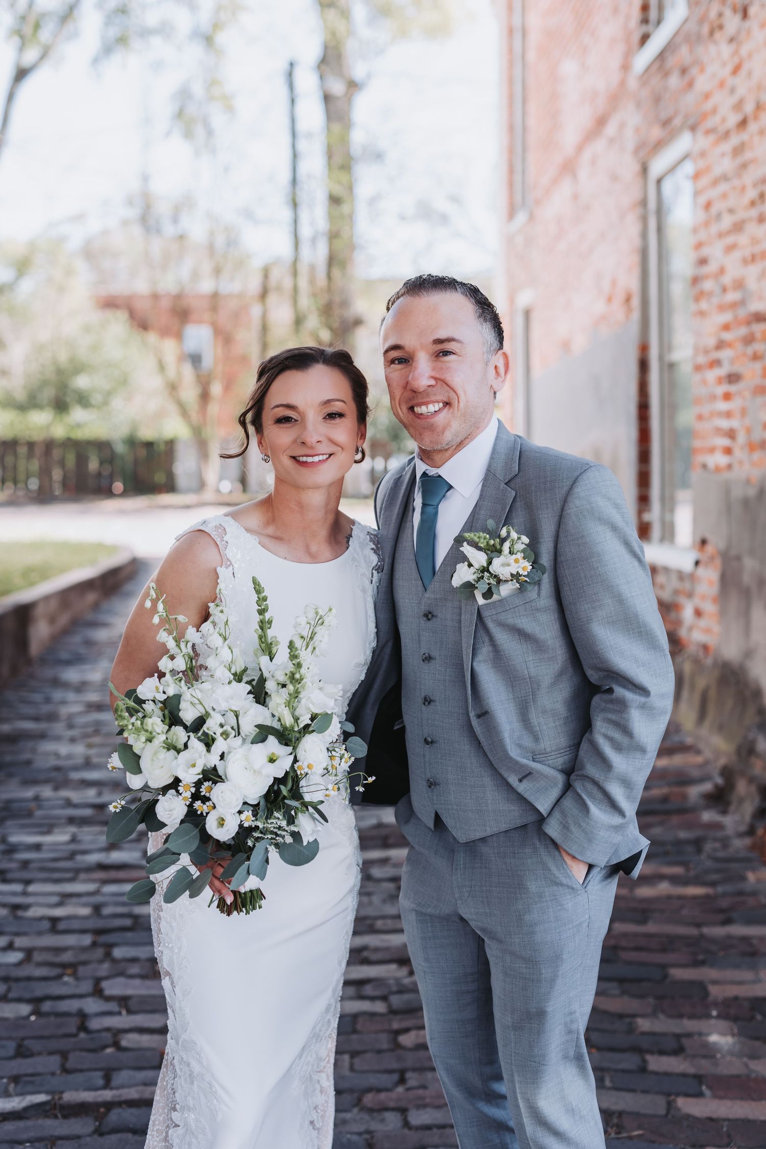 Smiling bride in white lace gown holds white floral bouquet beside groom in grey suit on cobblestone path.