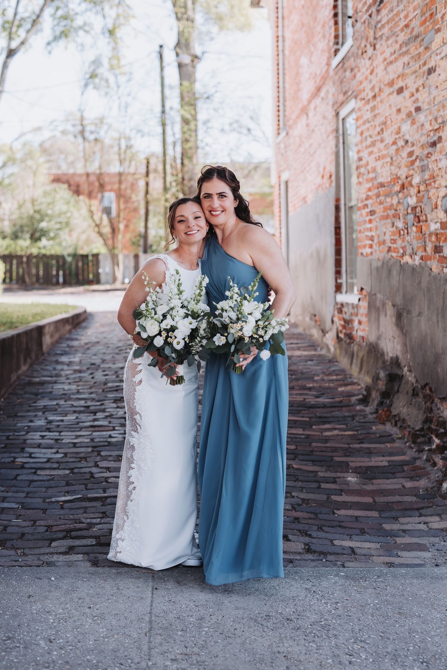 Bride and bridesmaid in blue gown smiling together holding white bouquets on cobblestone path.