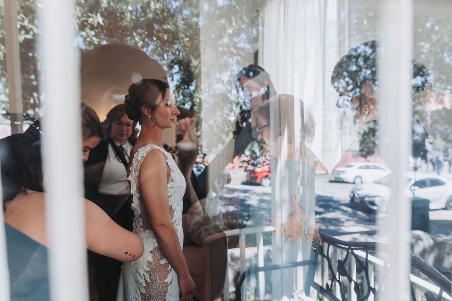 Bride in beaded gown viewed through window reflection surrounded by wedding guests indoors.