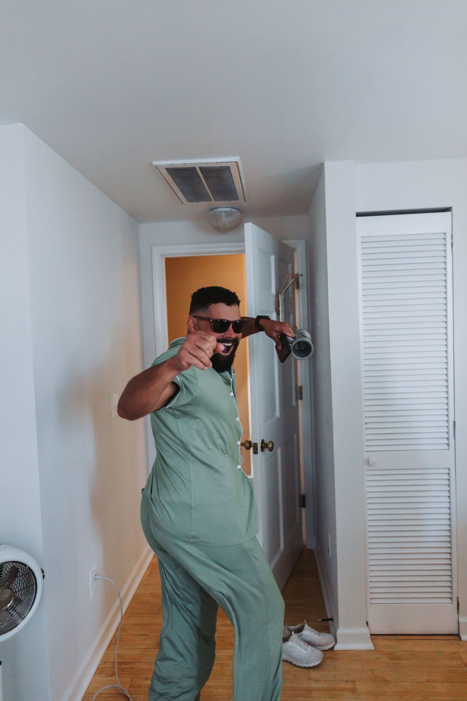 Man in sage green jumpsuit and sunglasses playfully poses with binoculars indoors at wedding venue.