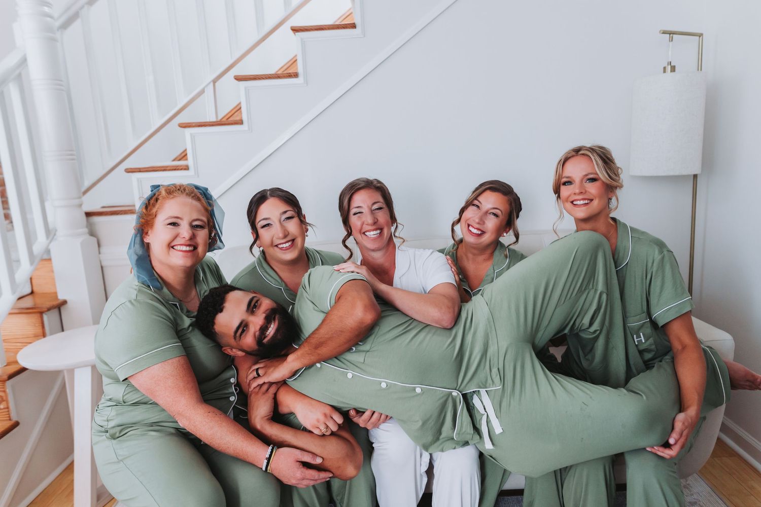Bride in white robe laughing with bridesmaids in sage green robes sitting on staircase.