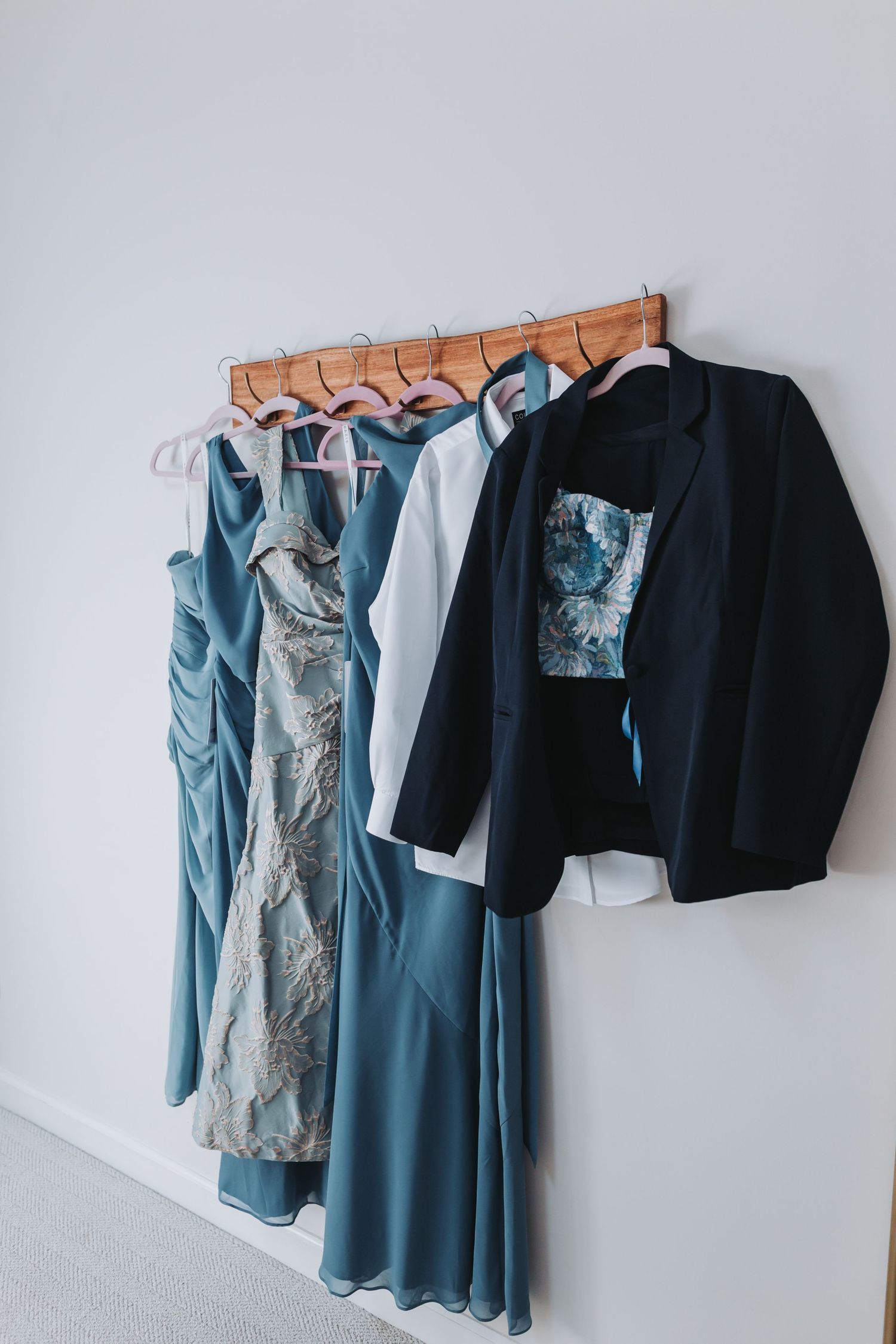 Bridesmaid dresses and groomsmen jacket hanging on wooden rack against white wall.