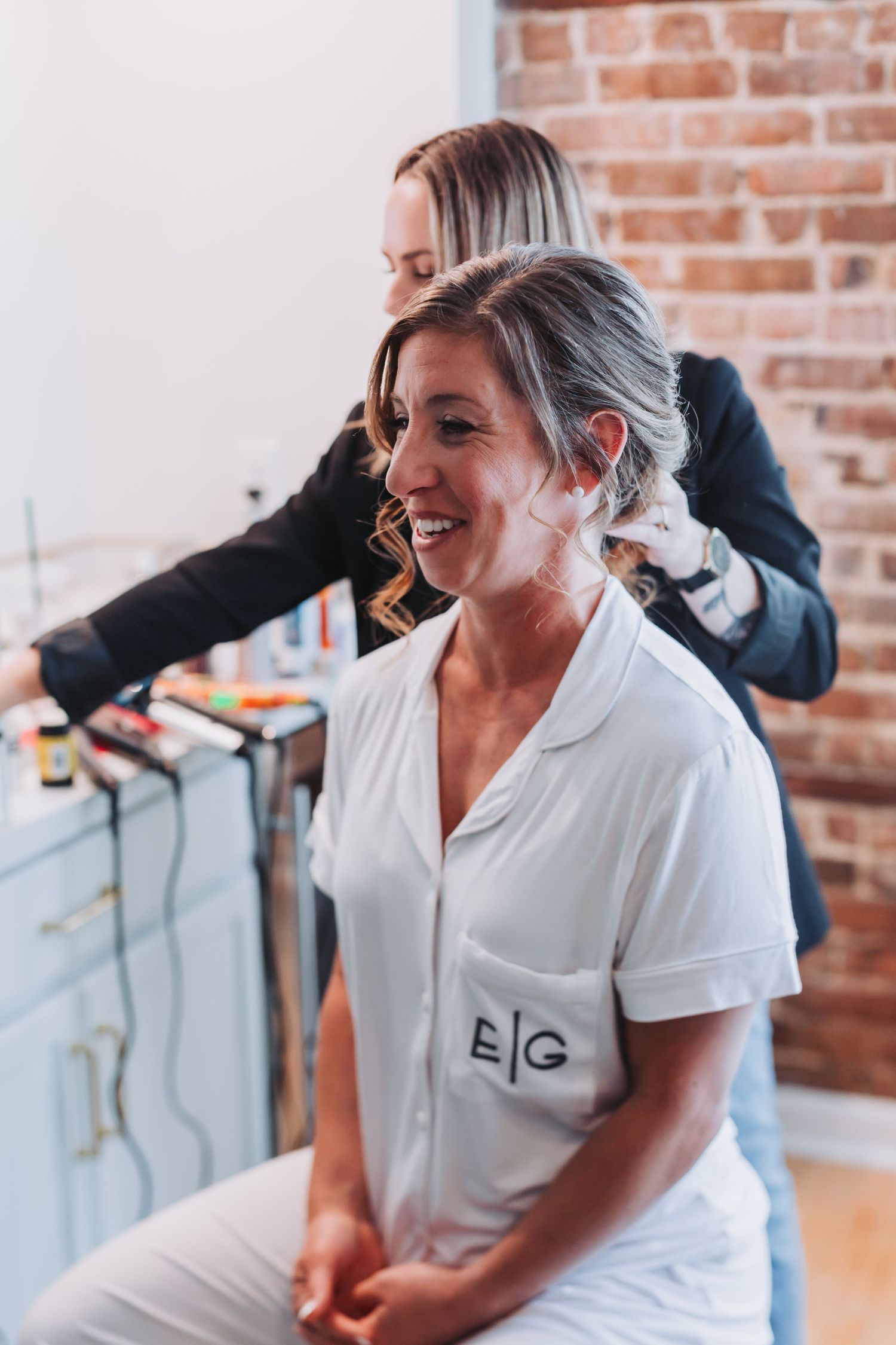 Bride smiling in white monogrammed pajamas while hairstylist works on her updo.