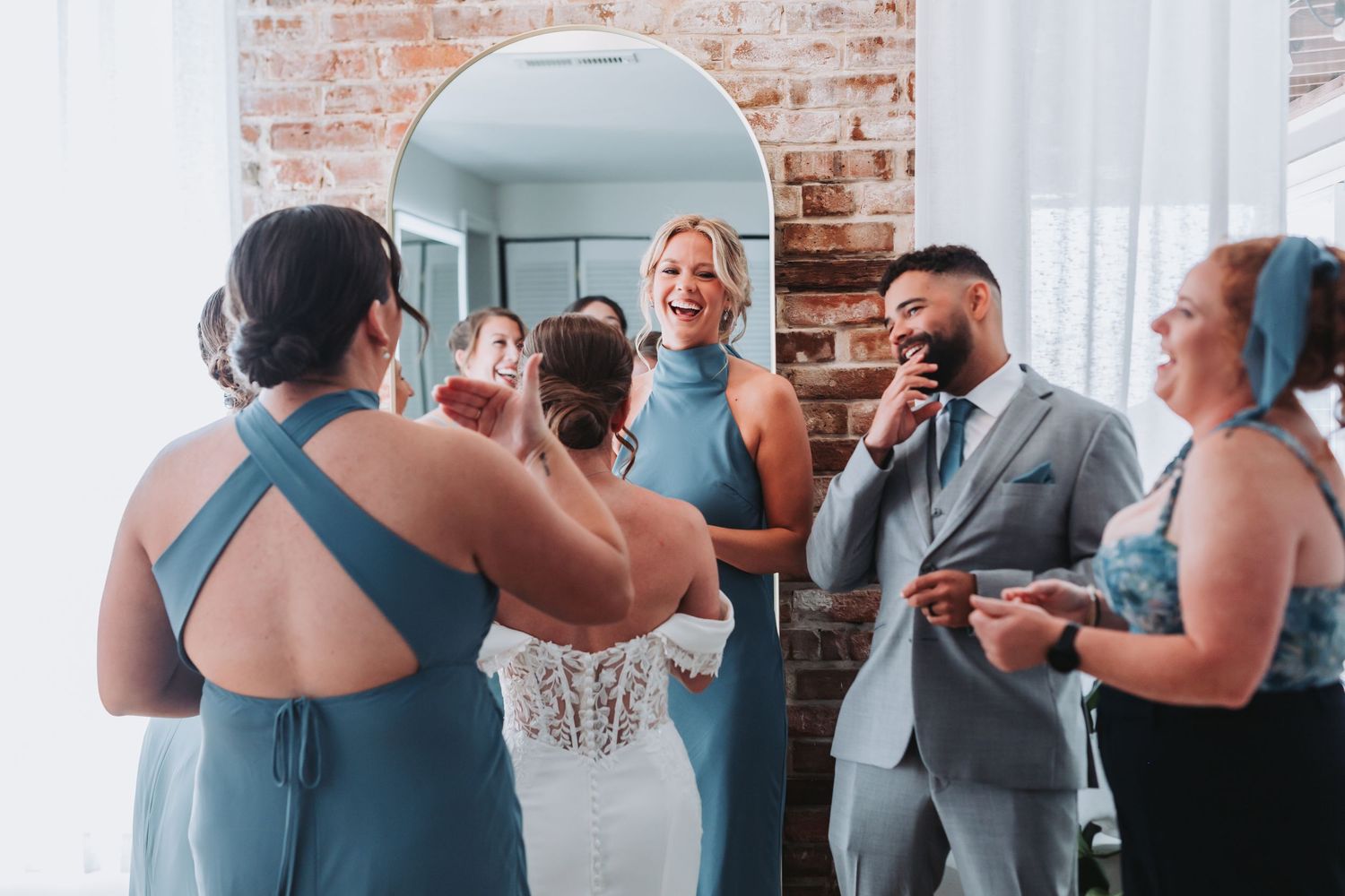 Bride in lace gown laughs with bridesmaids in dusty blue dresses and groomsman in gray suit near arched mirror.
